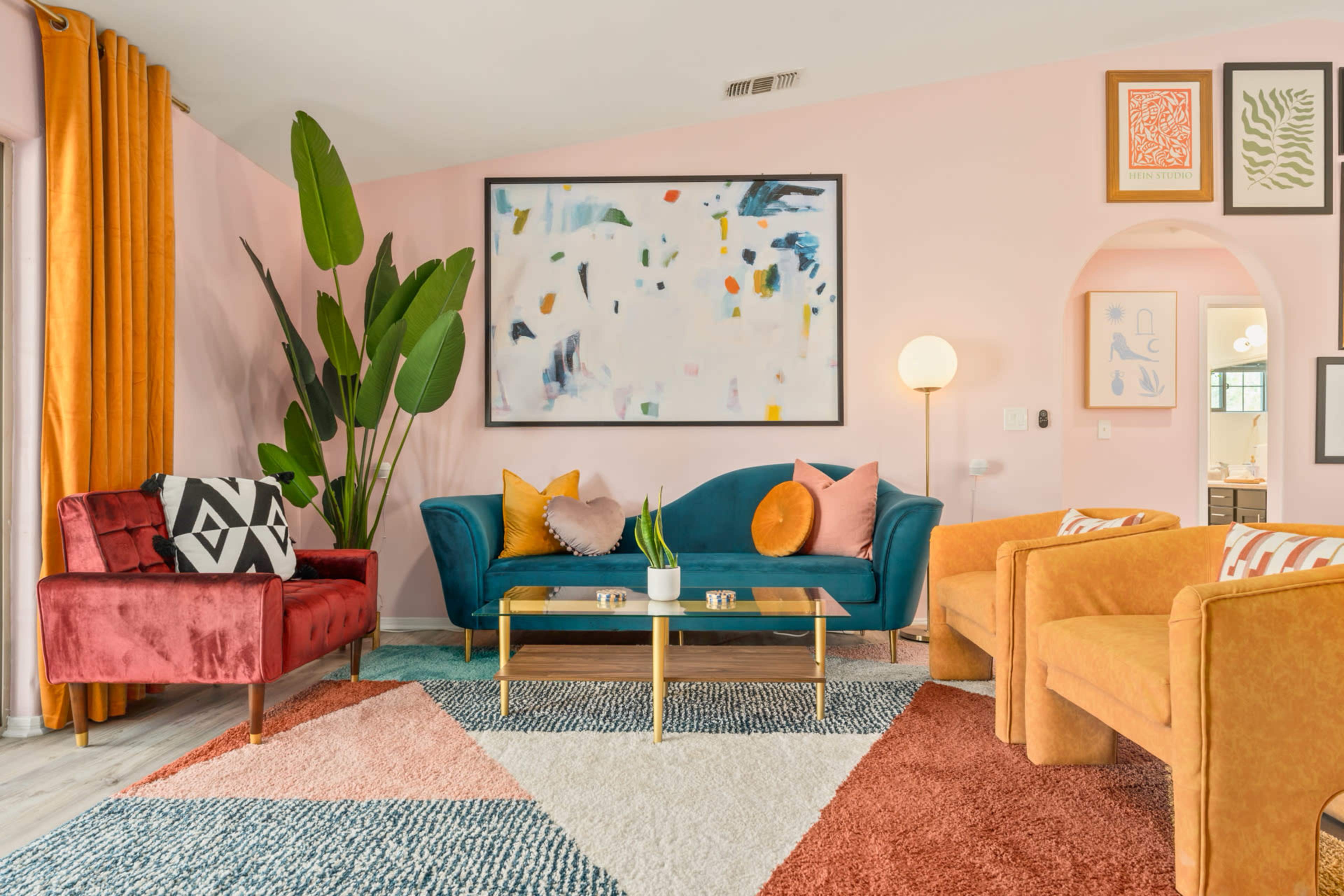A brightly colored living room with a pink wall, a large abstract painting, and a mix of furniture including a teal sofa, orange chairs, and a red velvet loveseat arranged on a patterned rug.