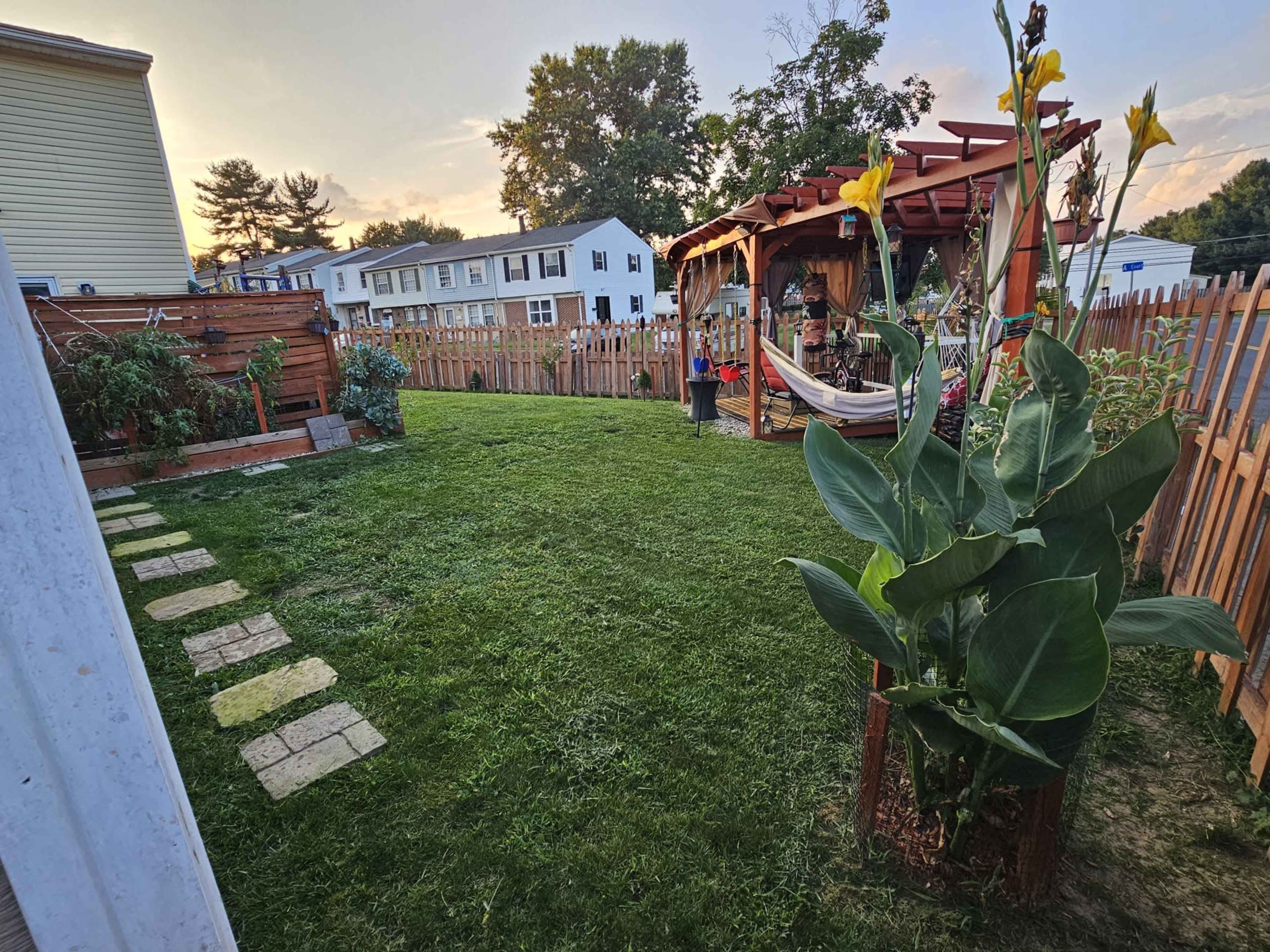 The image shows a landscaped backyard with a grassy area, a wooden gazebo, and a flower garden near a fenced property.