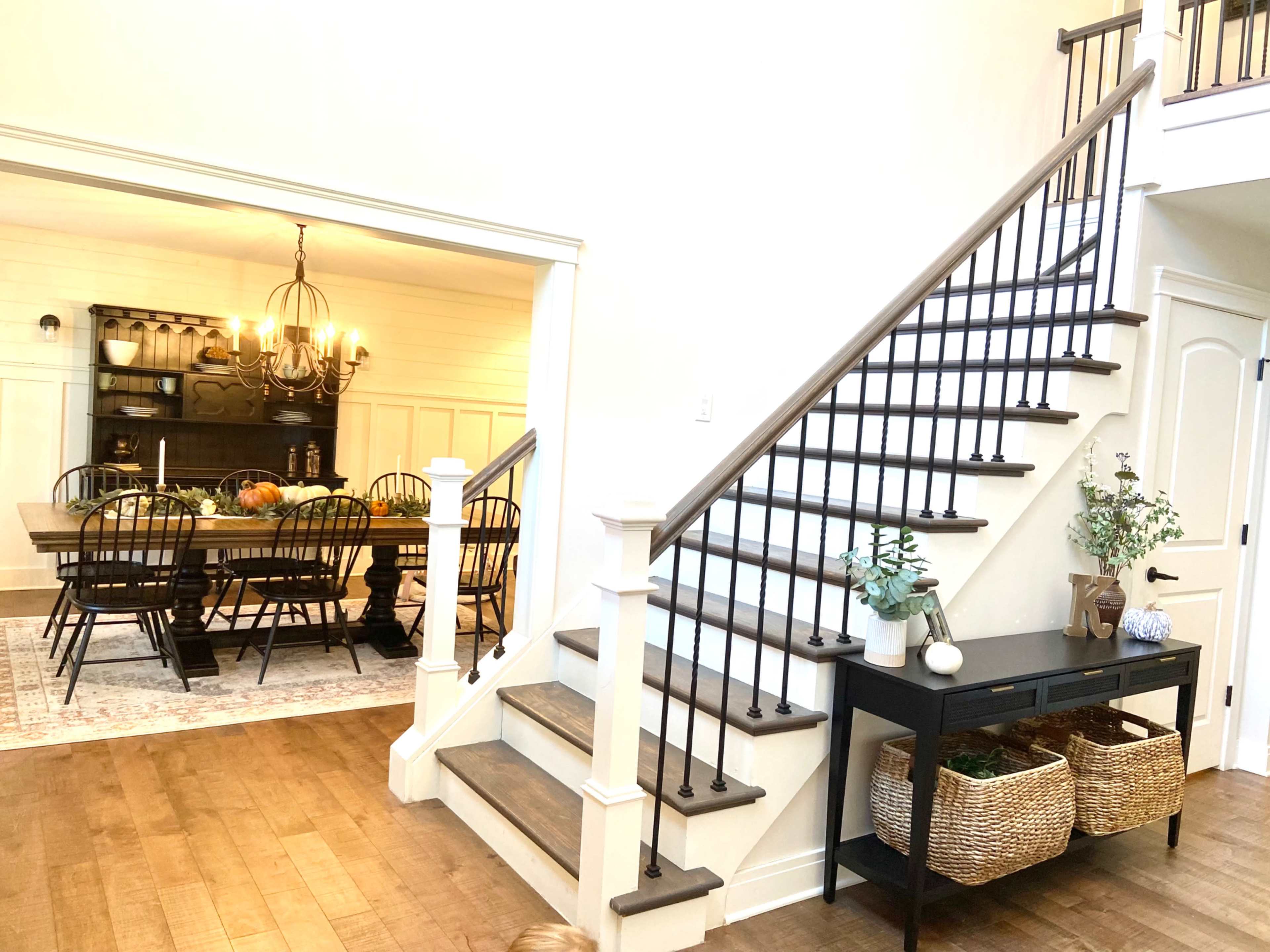 A staircase with a wooden handrail leads to an upper level, while a dining area with a long table and chairs is visible through an open doorway beside it.