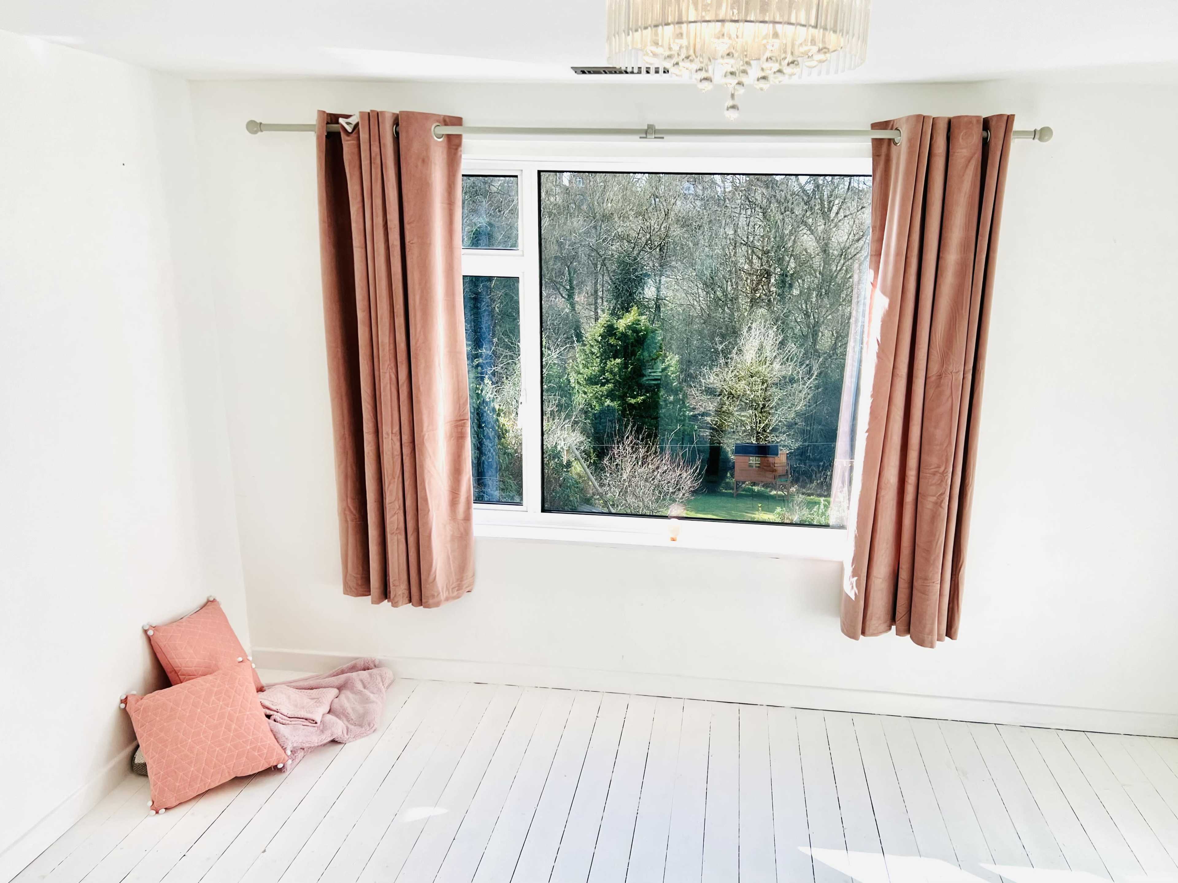 A bright, empty room with white walls, a large window featuring peach curtains, and two cushions on the floor.