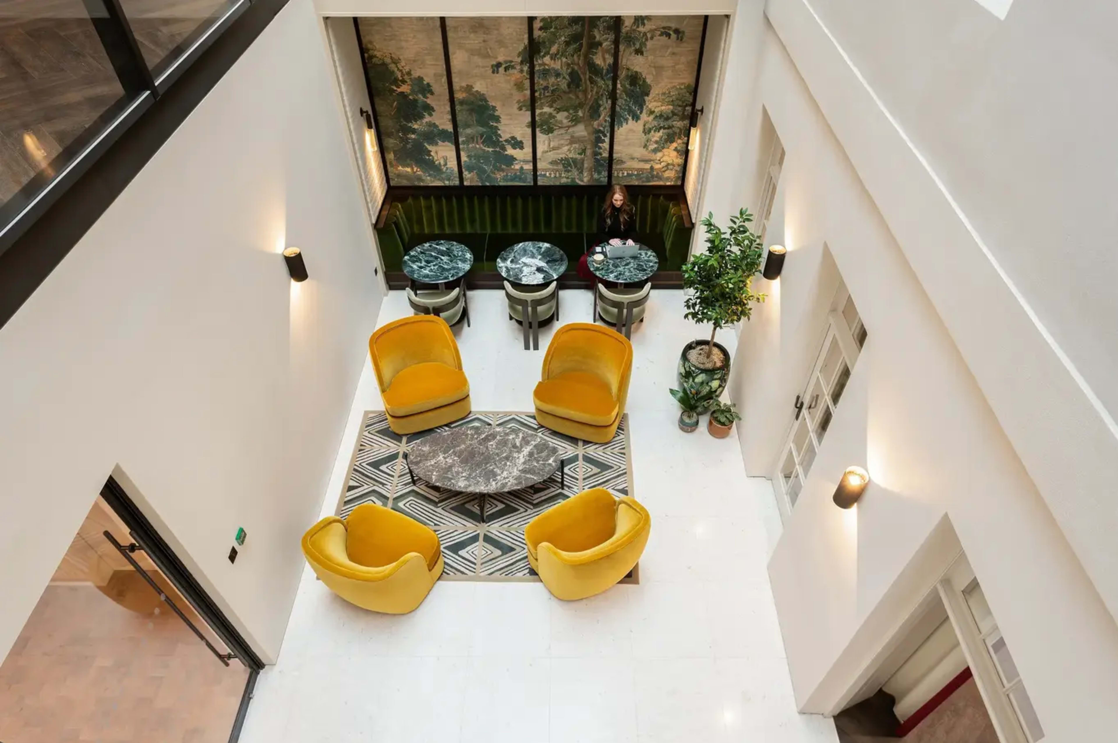 The image shows a spacious interior with yellow armchairs, marble-top tables, and large windows featuring a mural, all arranged around a patterned rug.