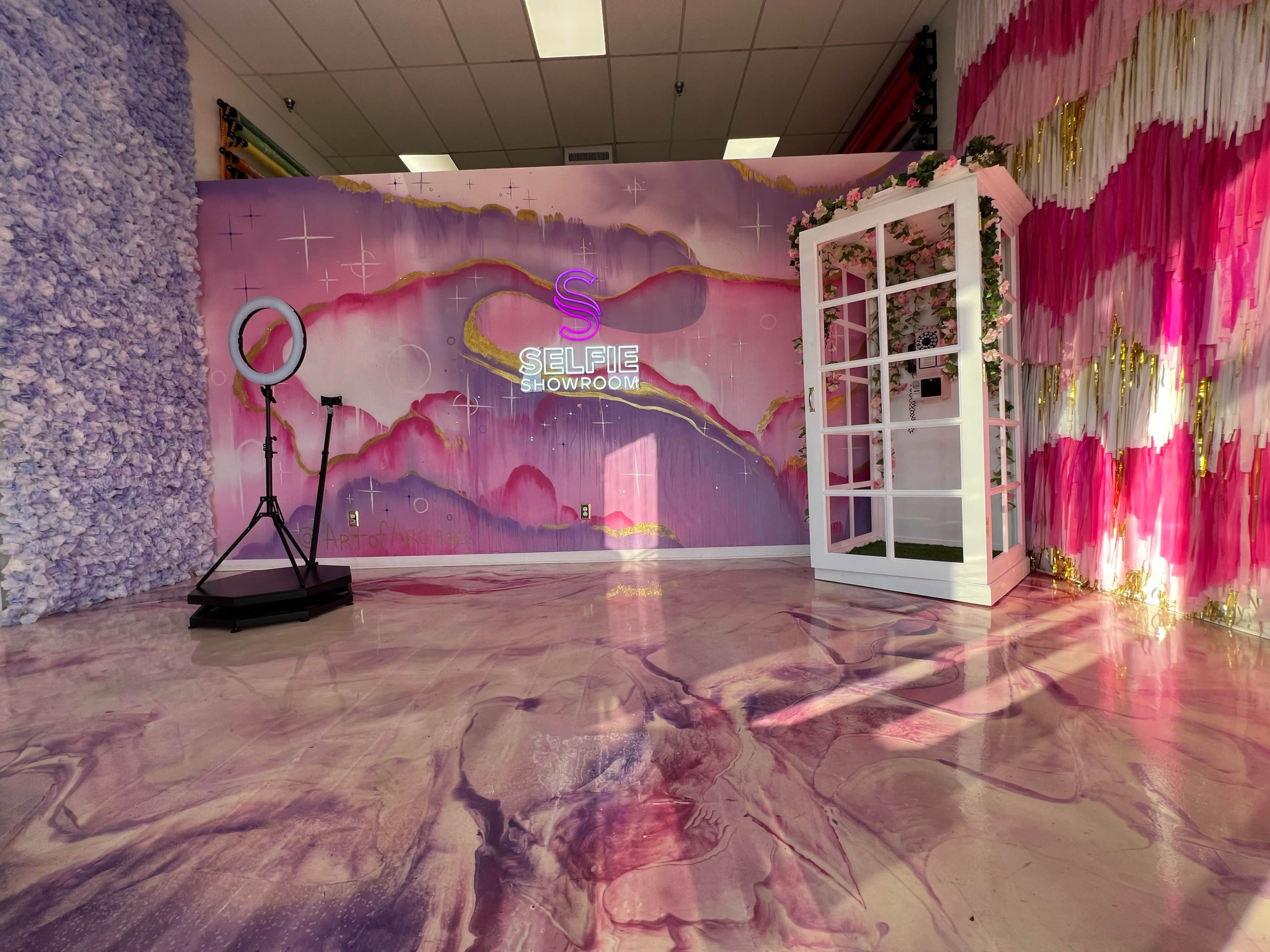 The image shows a brightly decorated showroom with a colorful wall backdrop, a ring light on a stand, and a white, glass-paneled photo booth.