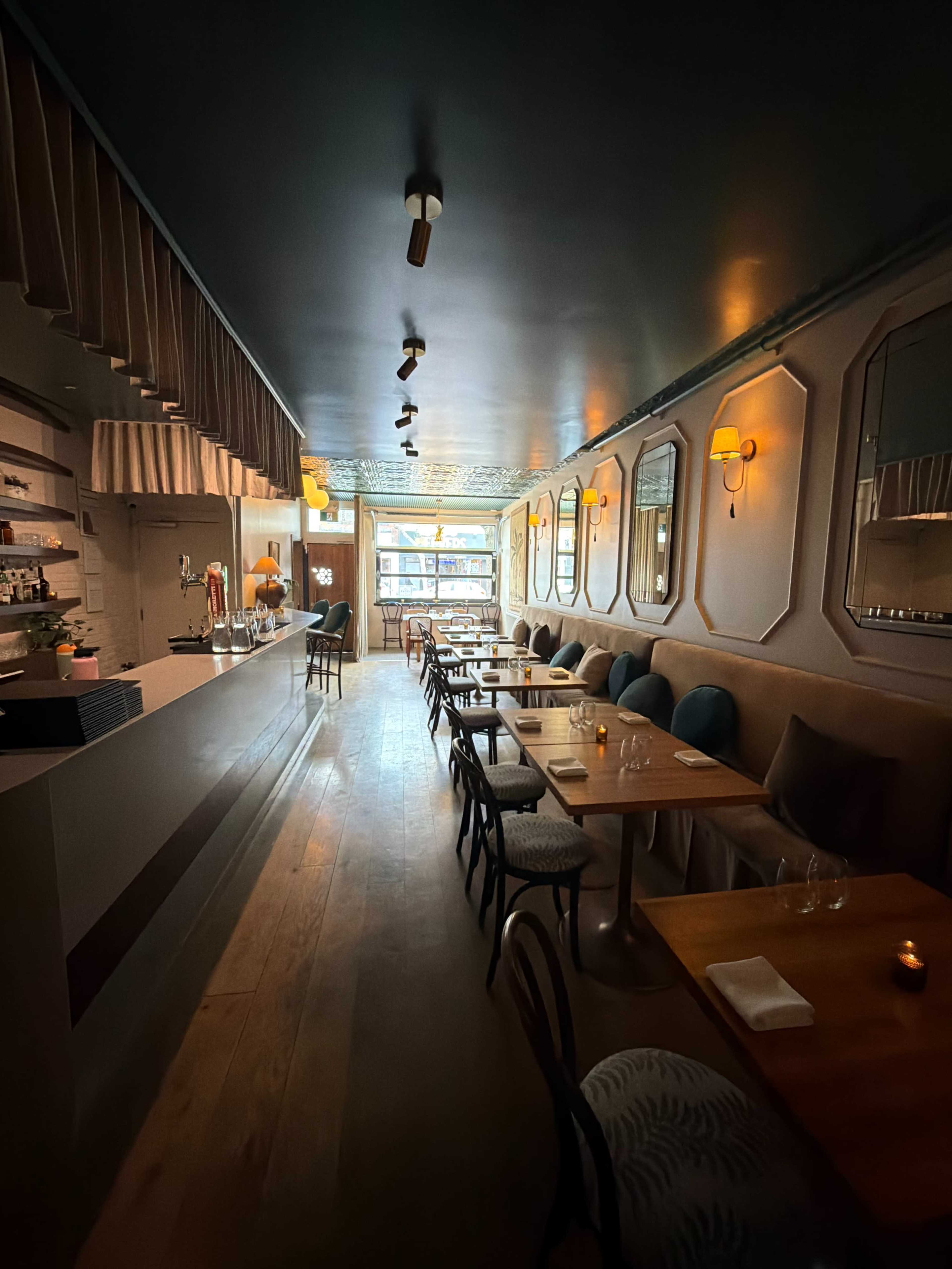 The image shows a long, narrow restaurant interior featuring a bar on one side and tables with chairs arranged along the other side, illuminated by soft lighting.