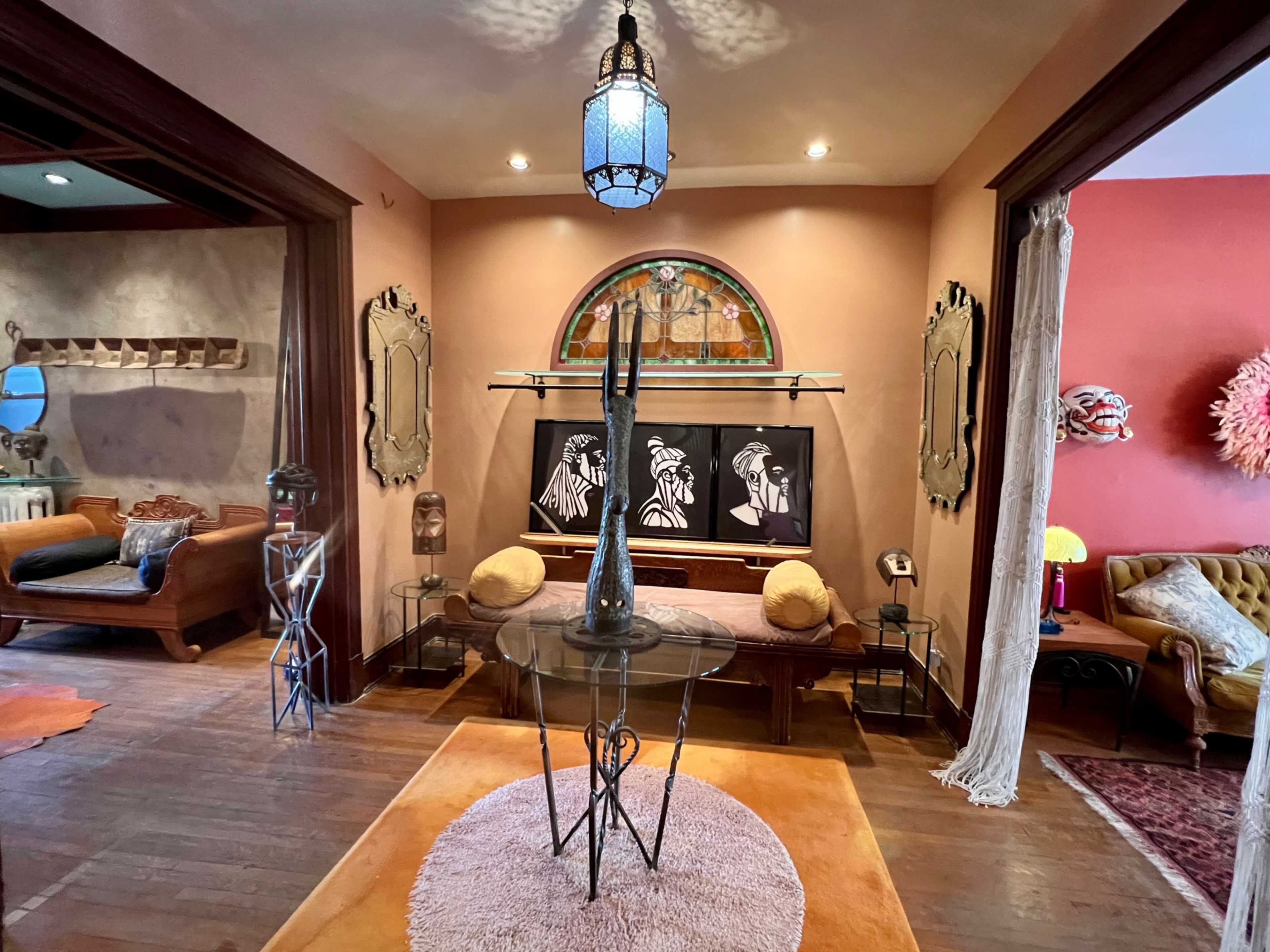 The scene depicts a stylish interior featuring a glass table with a tall vase, framed artwork on the wall, and various decorative elements in a warm, inviting room.