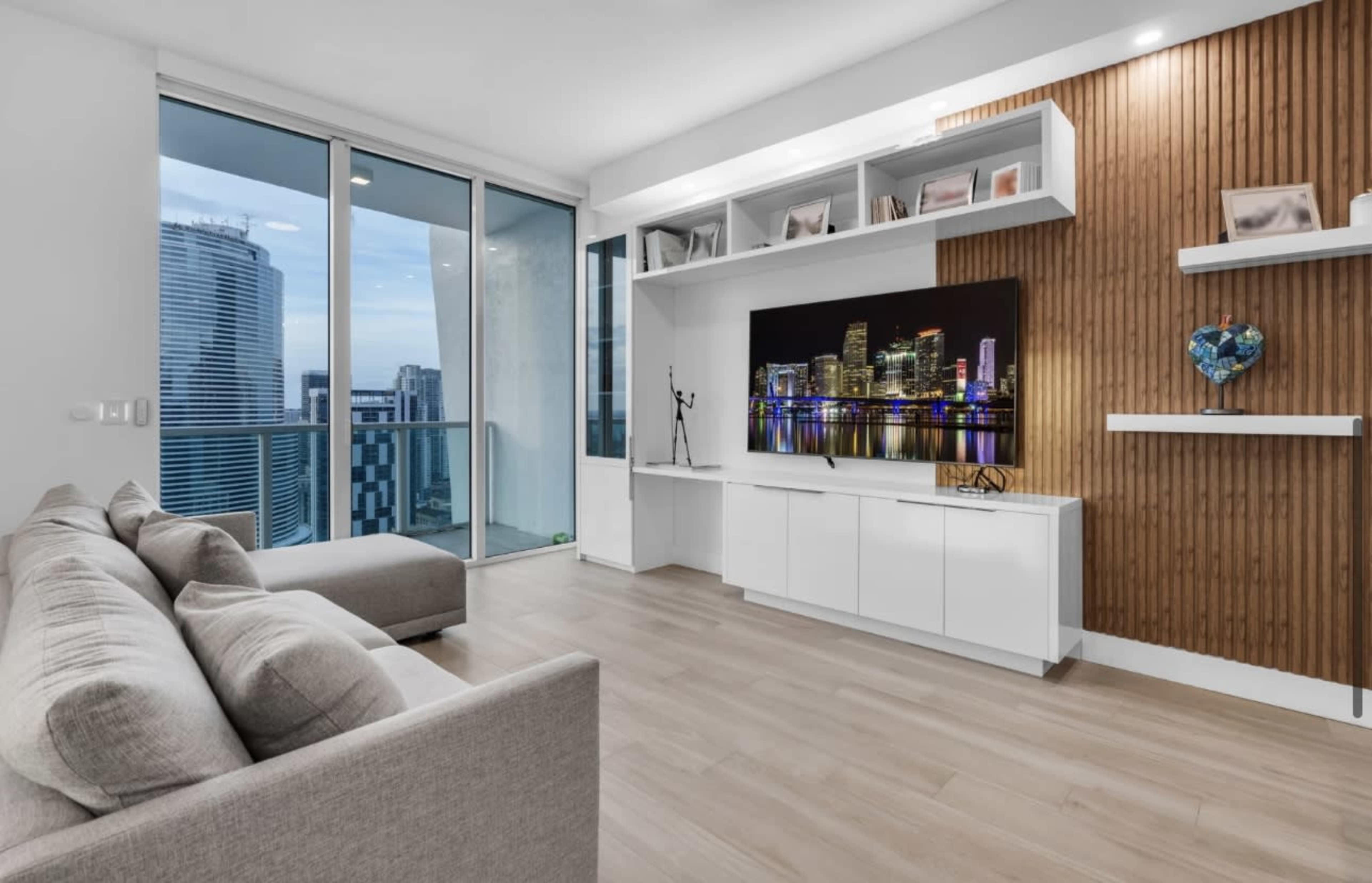 A modern living room features a beige sofa, a wall-mounted television, and wooden accents, overlooking a city skyline through large glass windows.