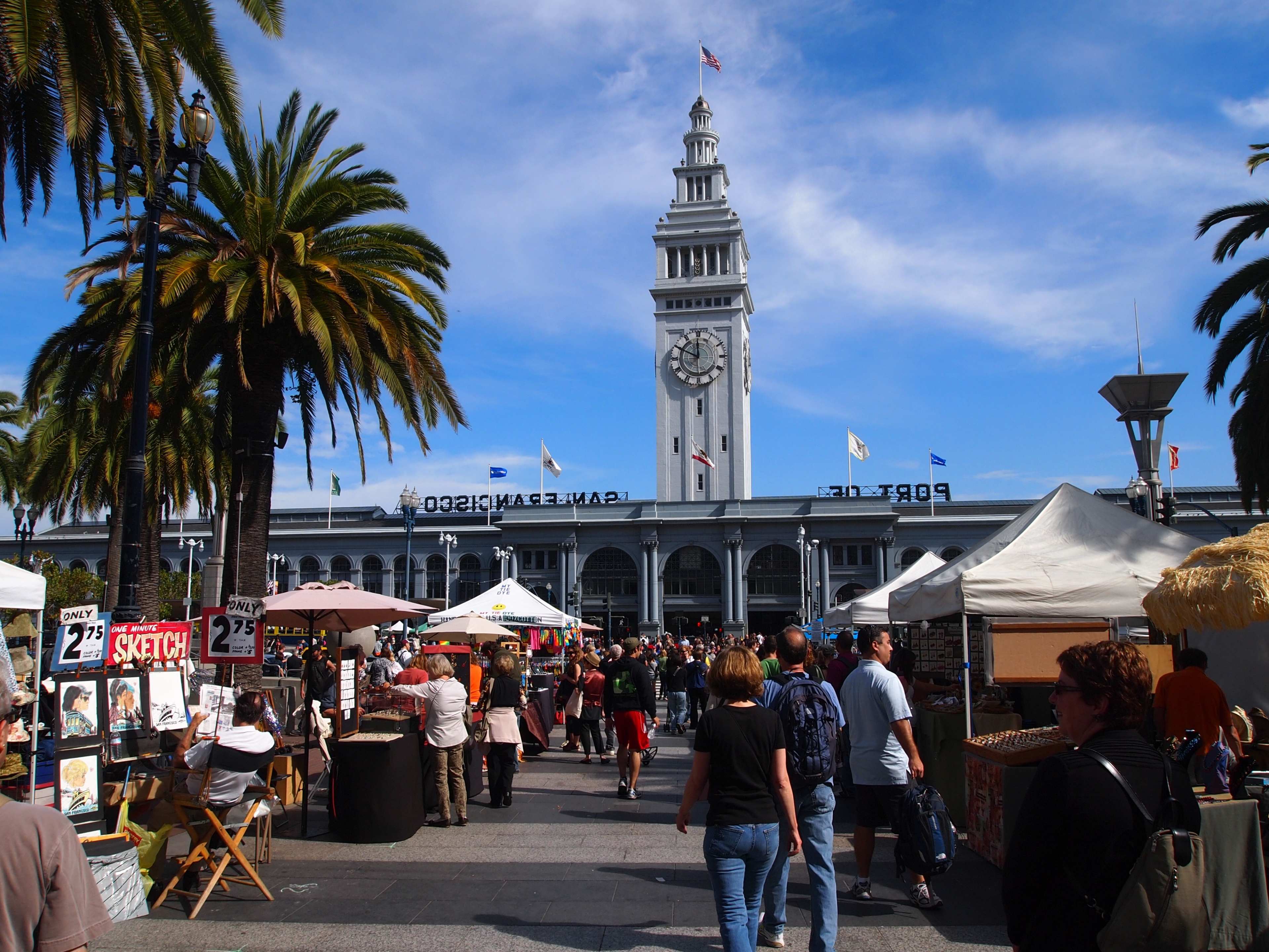 A bustling waterfront market scene is depicted with people walking along a promenade beside palm trees, featuring various stalls and a tall clock tower in the background.