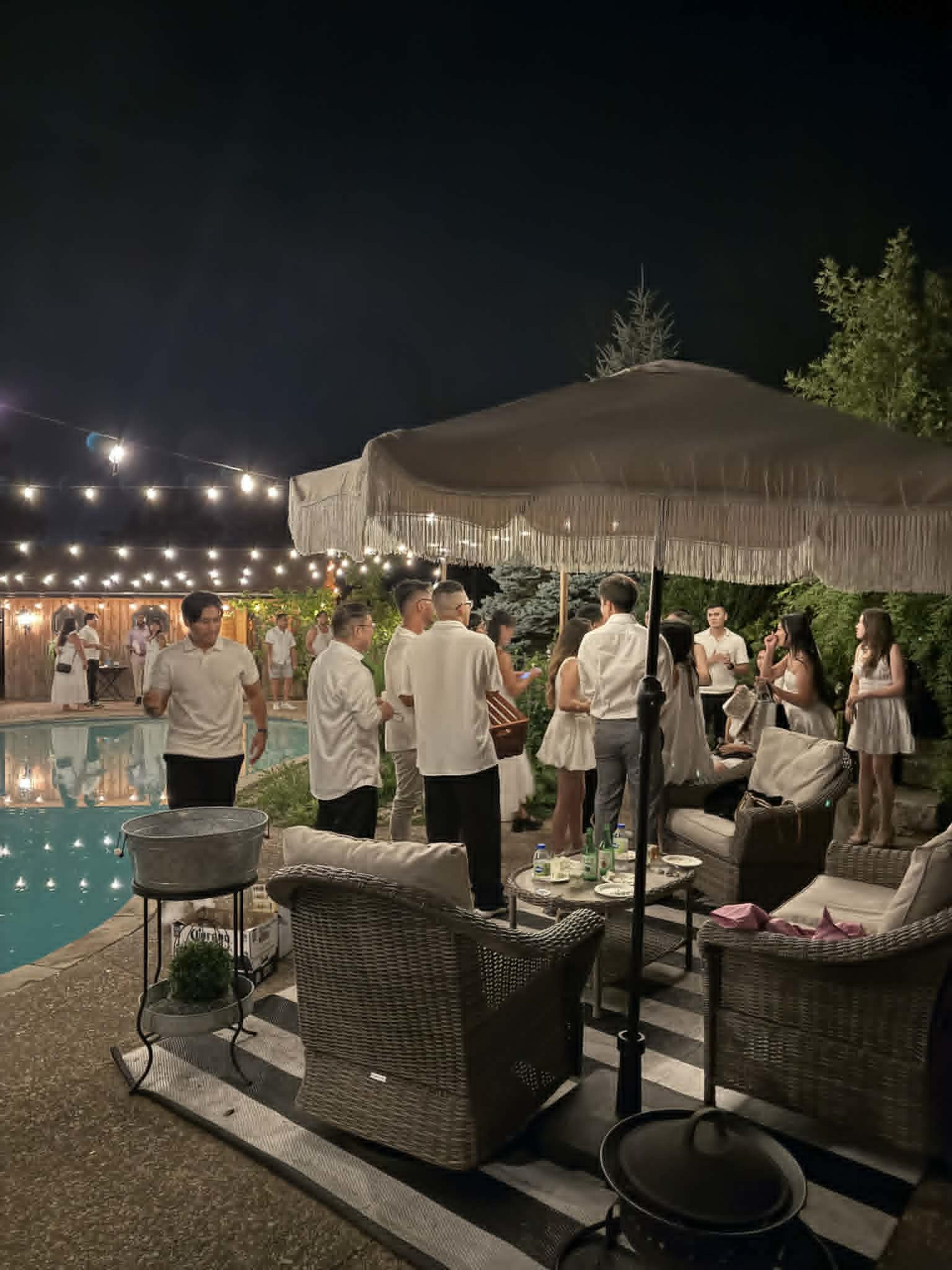 A group of people in white clothing socialize by a pool surrounded by string lights at night.