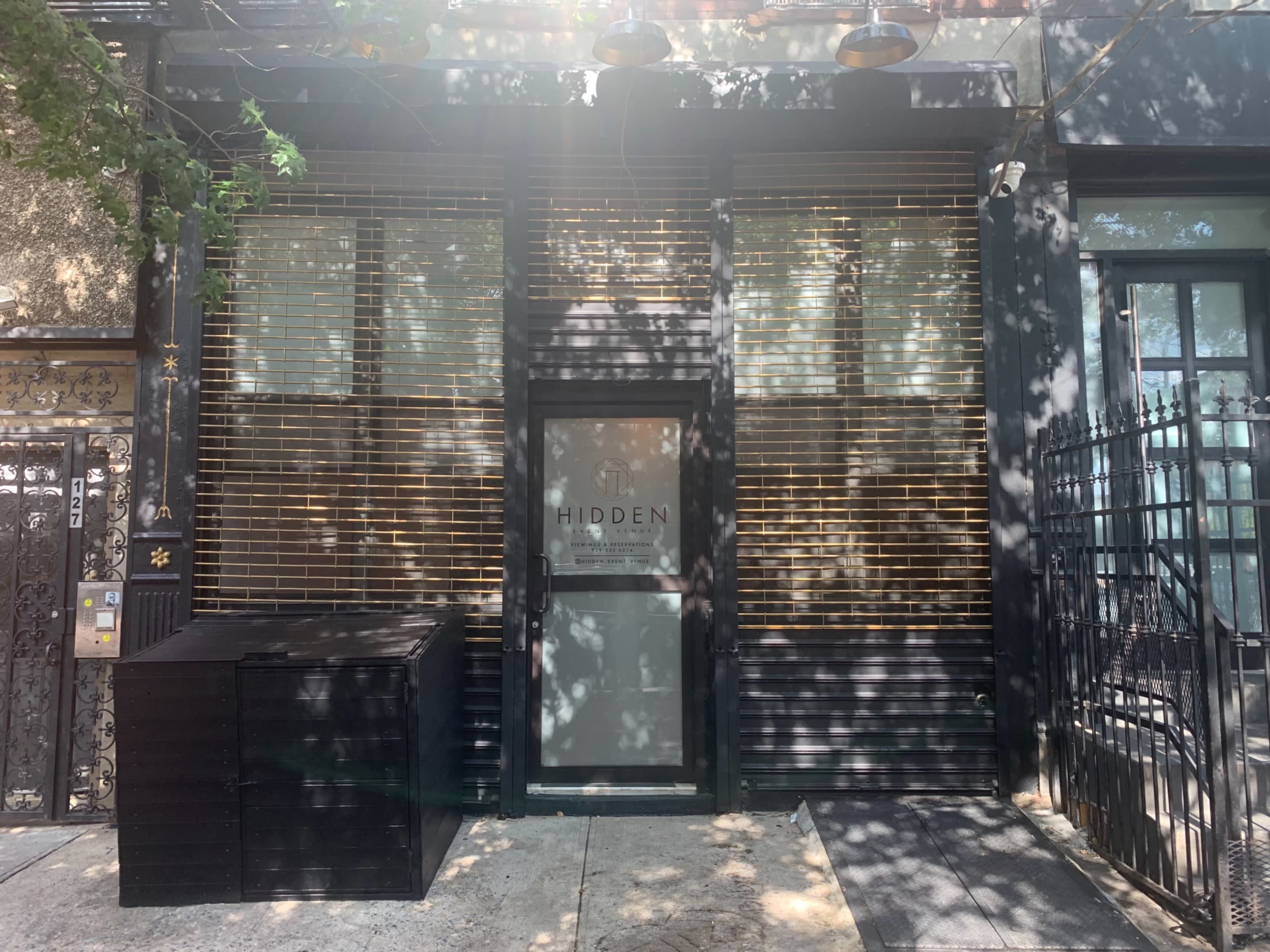 The front entrance of a building with large glass windows and a dark facade, featuring a sign that says "HIDDEN."