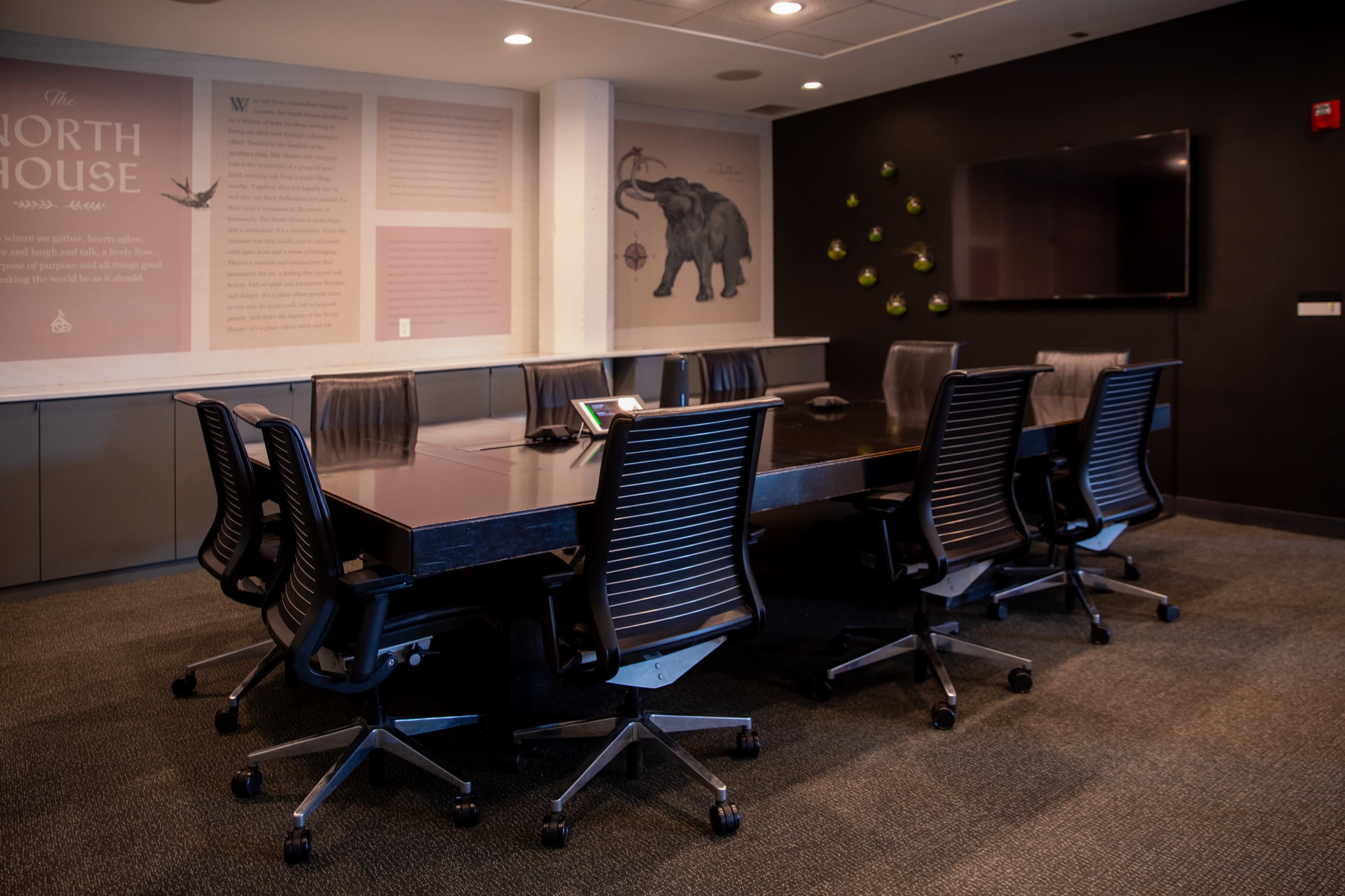 A modern conference room features a large rectangular table surrounded by ergonomic chairs, with a wall adorned by graphics and an elephant design.