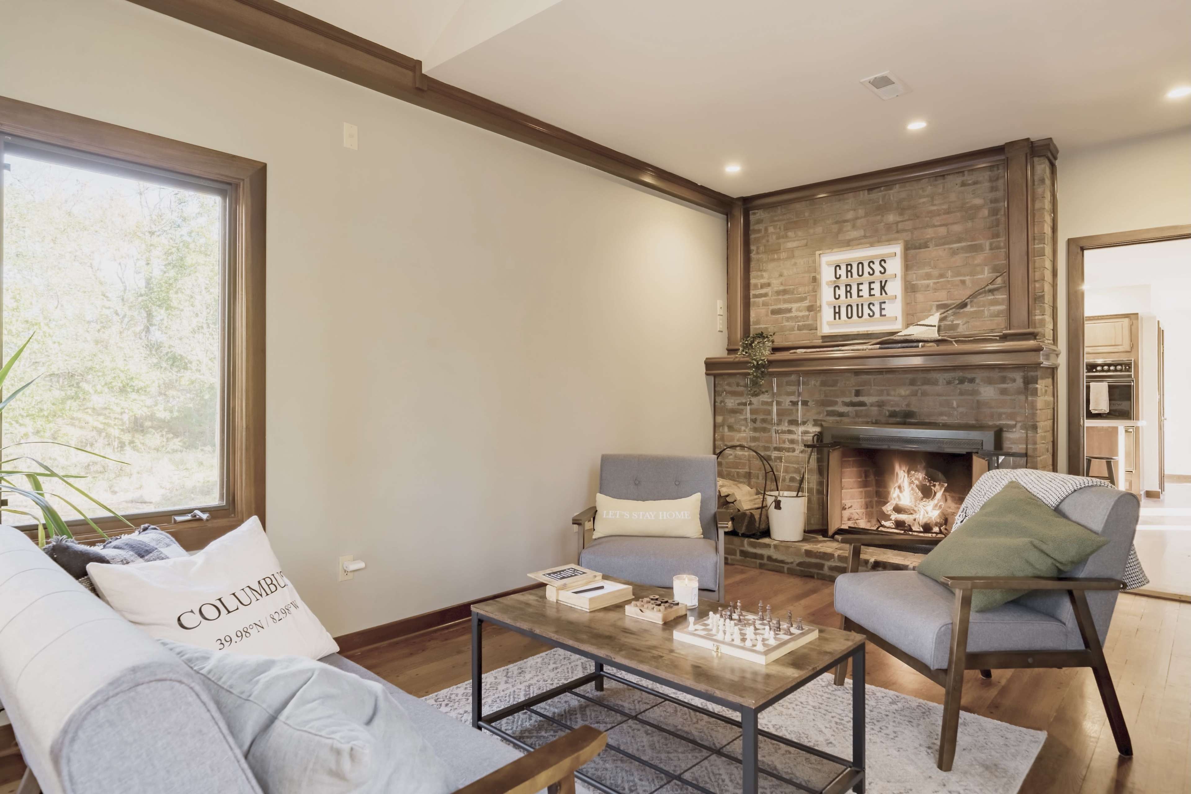 A cozy living room with a brick fireplace, two comfortable chairs, a wooden coffee table, and a large window letting in natural light.