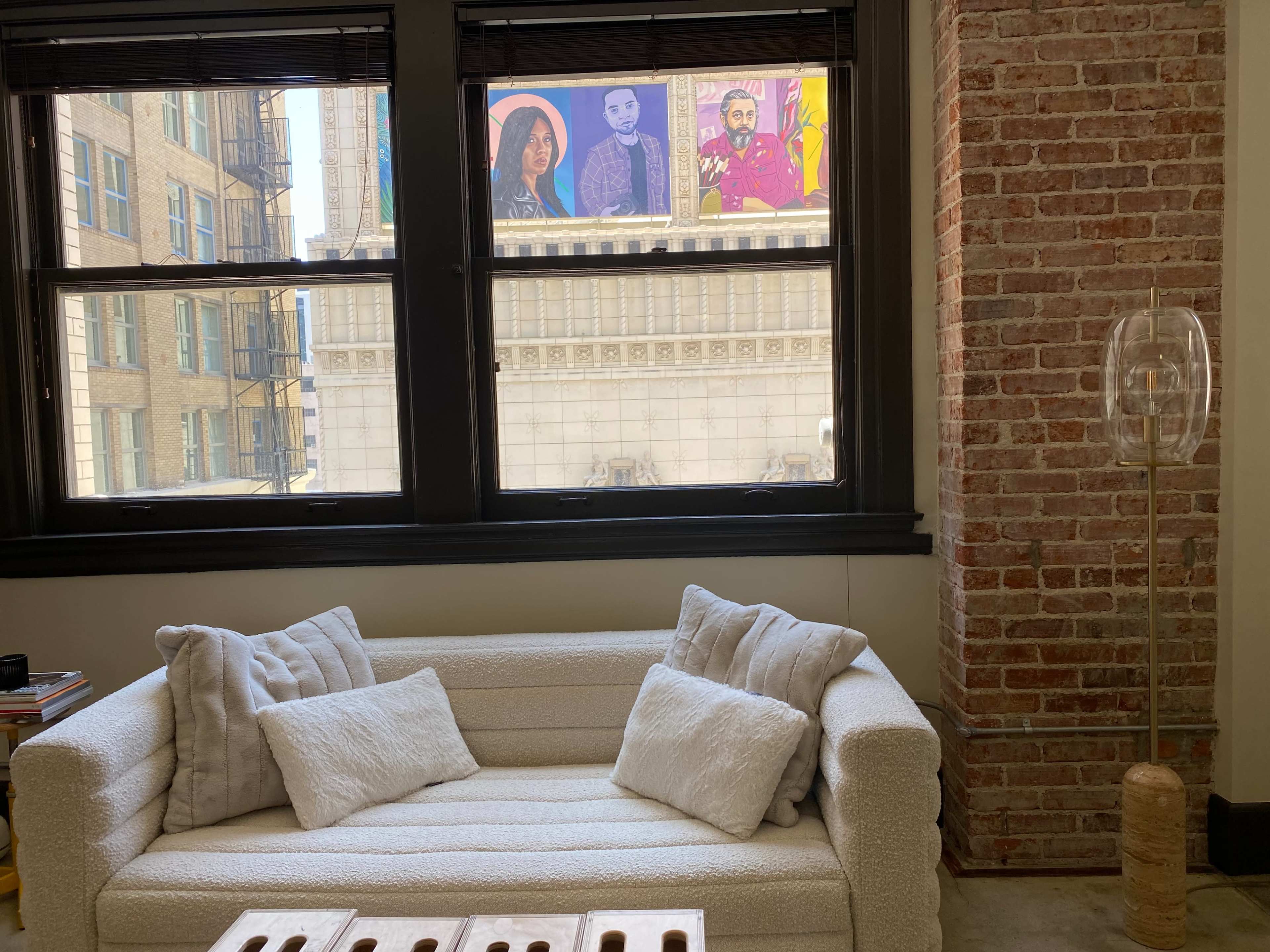 A white, upholstered couch with decorative pillows sits in front of a large window showcasing colorful street art on a building across the street.