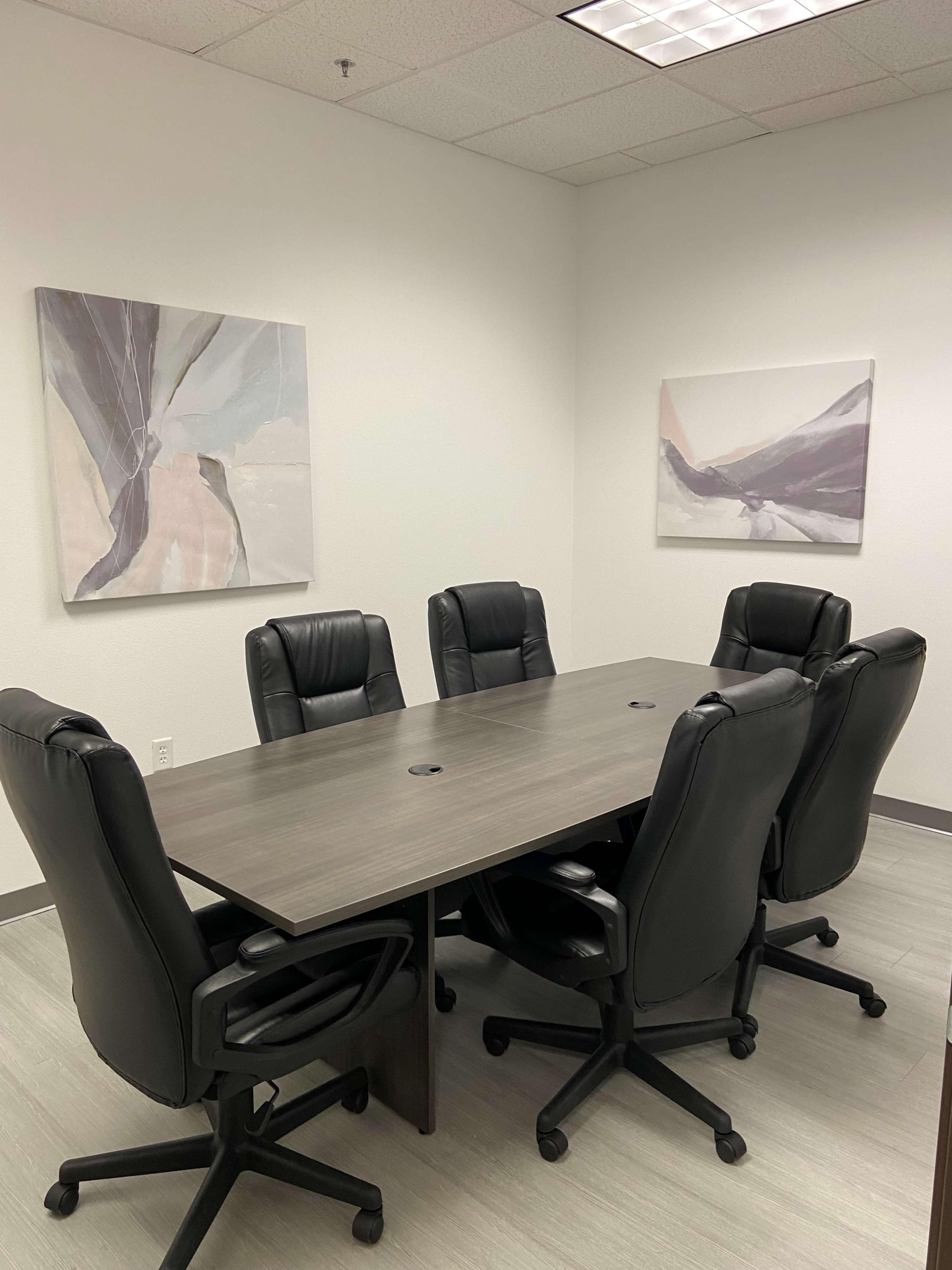 A modern conference room features a large table surrounded by six black rolling chairs, with abstract artwork hanging on the walls.