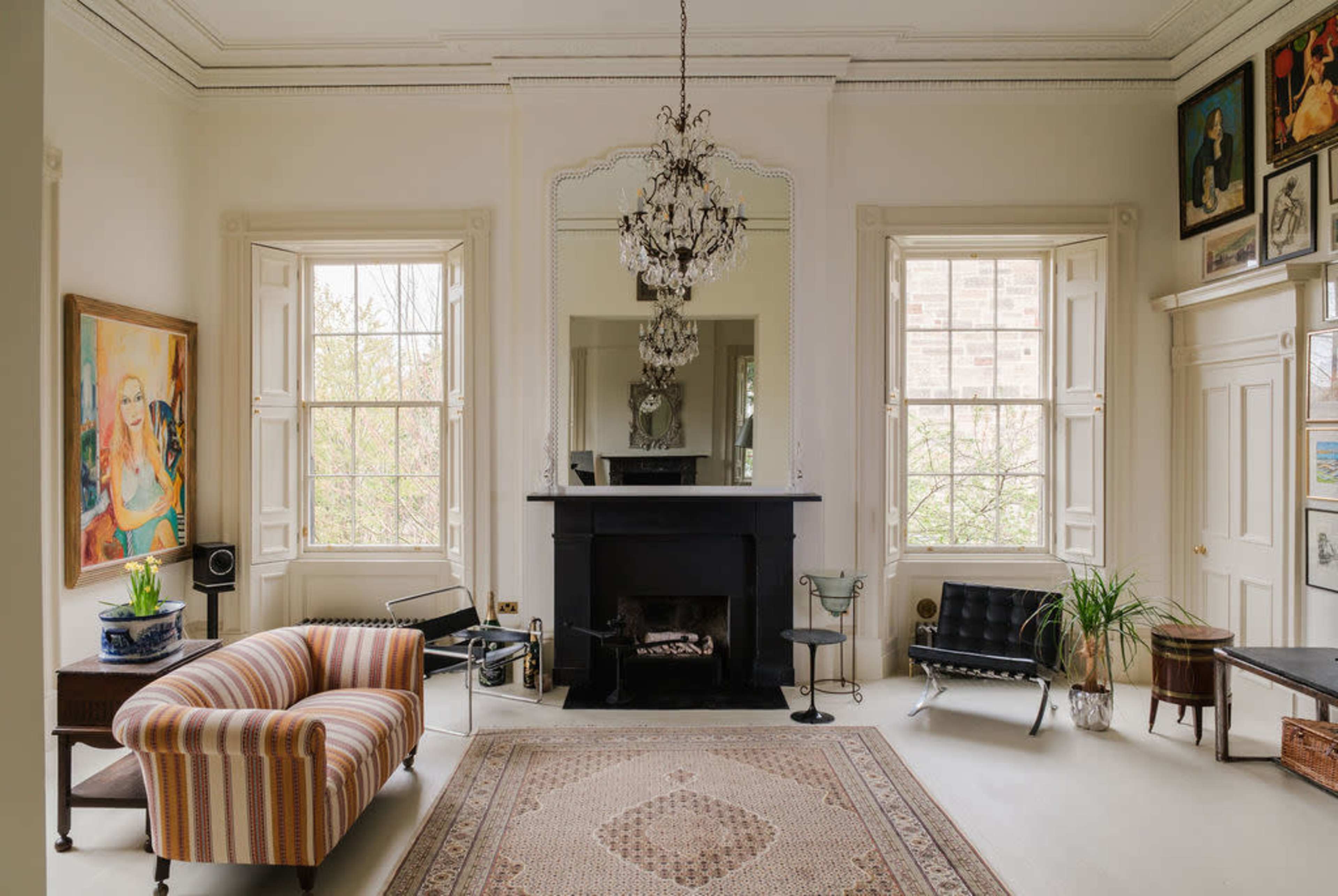 The image shows a spacious living room with large windows, a central chandelier, a black fireplace, and a mix of modern and vintage furniture.