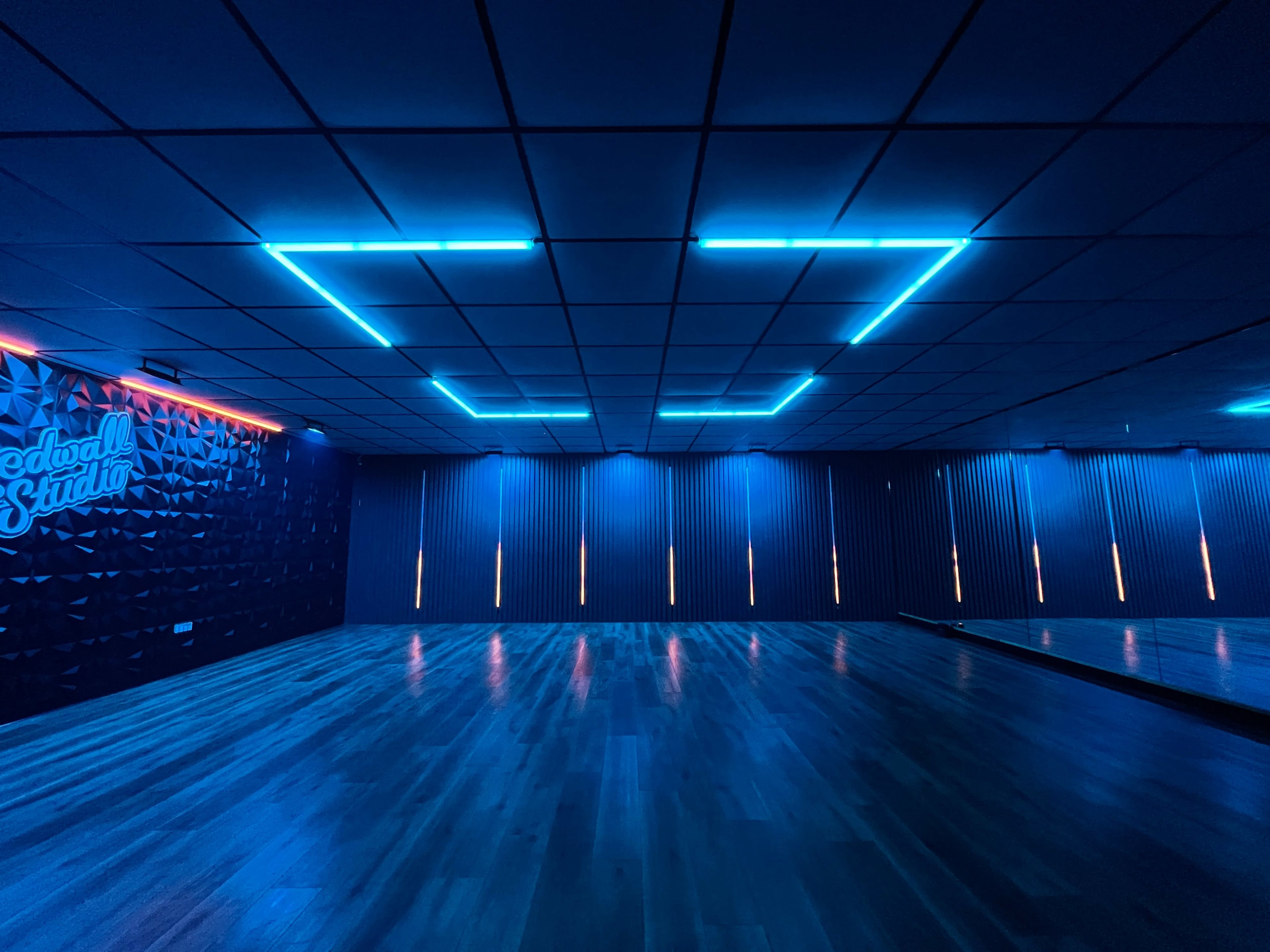 A dimly lit dance studio featuring blue neon lights along the ceiling and walls, with mirrored panels on one side.
