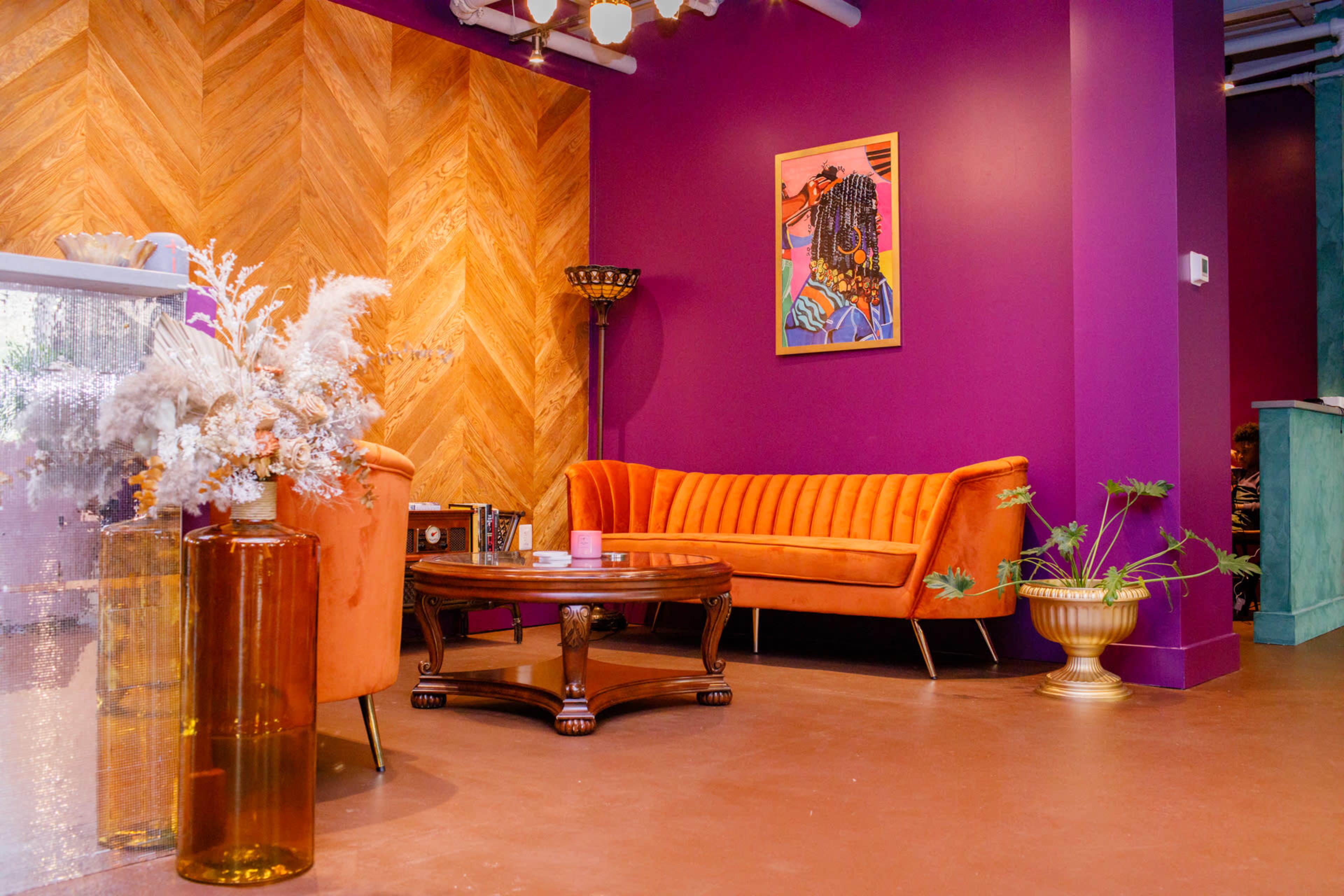 The image features a vibrant indoor lounge area with a purple wall, an orange velvet sofa, a wooden coffee table, and decorative plants.