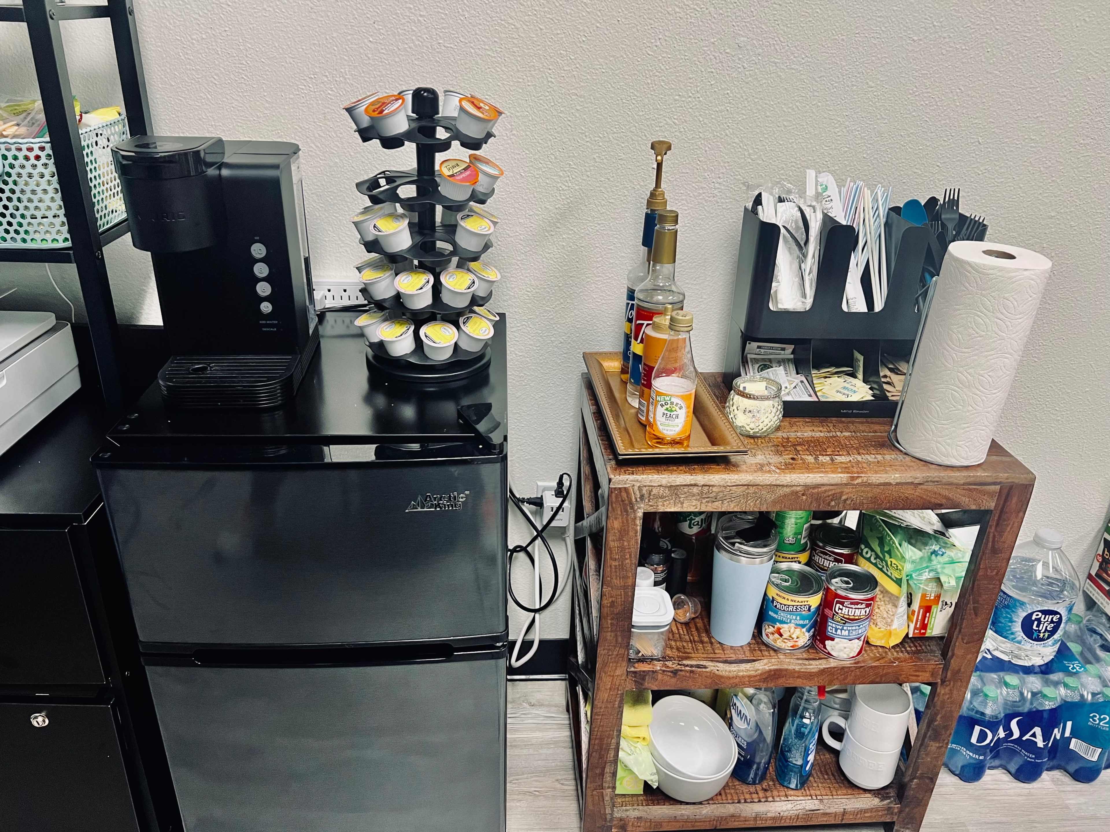A small kitchen area featuring a compact refrigerator, a coffee capsule holder, and a wooden cart filled with various kitchen items and supplies.