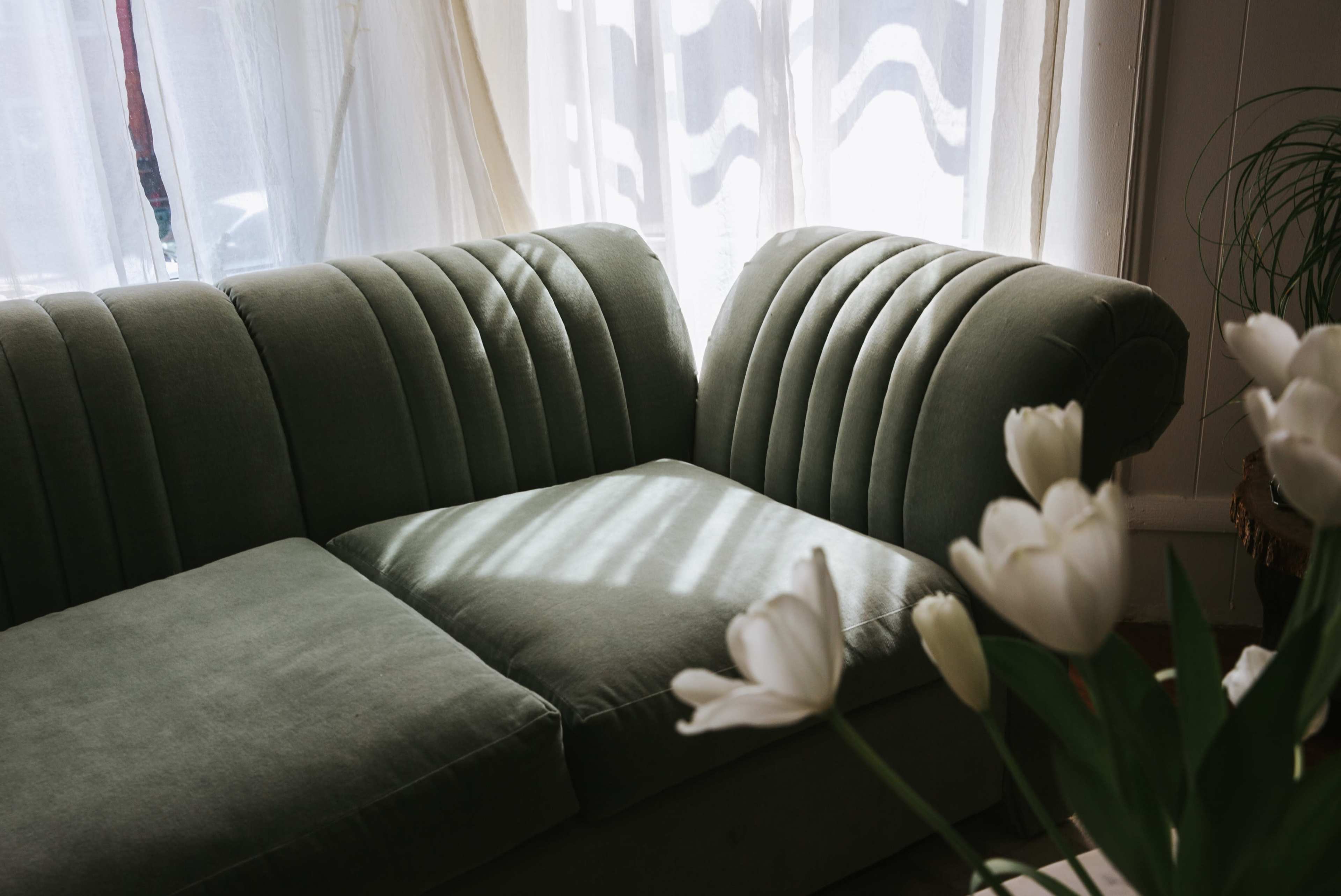 A green, ribbed sofa is positioned near a window, with light filtering through sheer curtains and a vase of white tulips in the foreground.