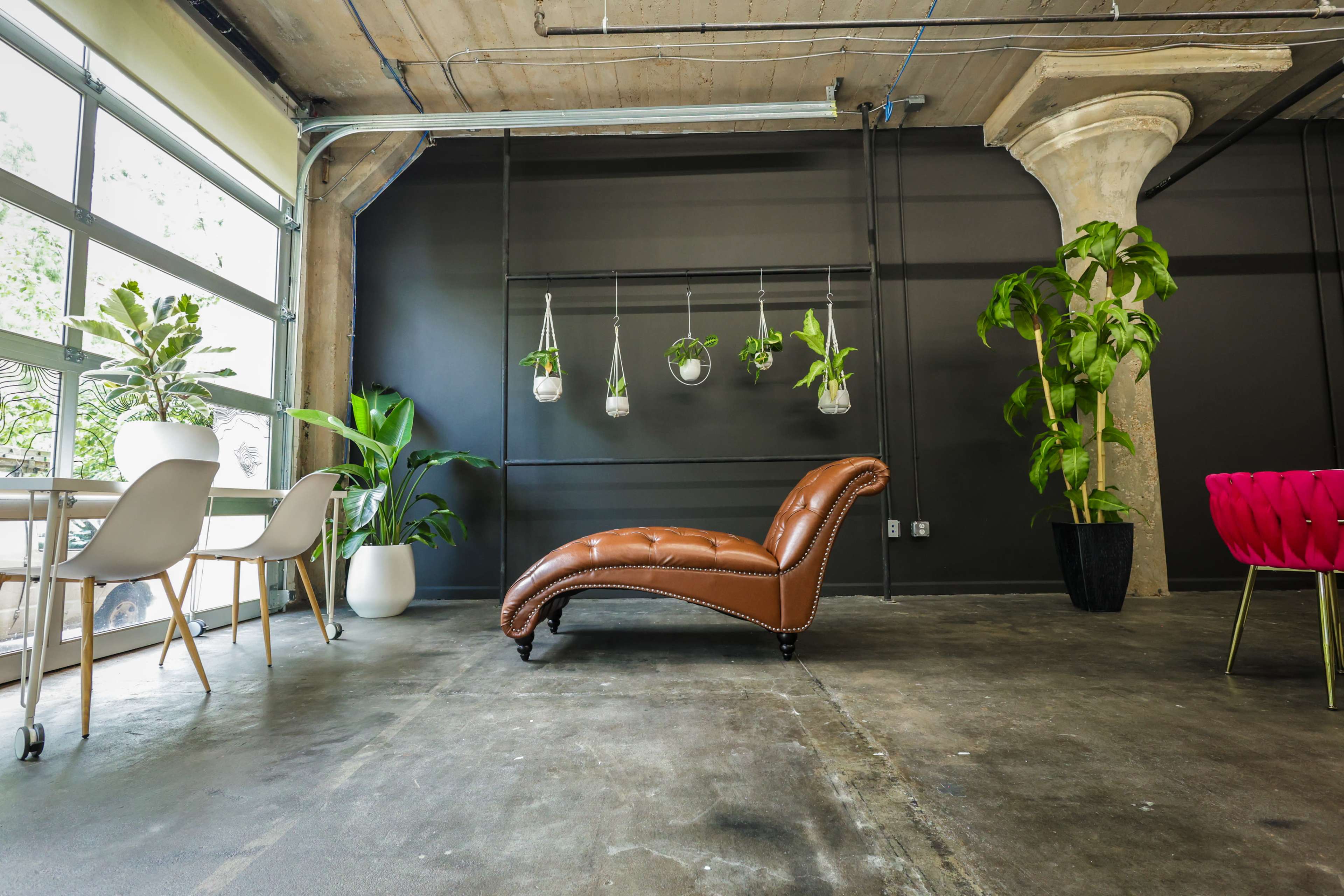 A modern interior space featuring a brown chaise lounge, a glass table with white chairs, hanging planters, and various plants against a dark wall and concrete floor.