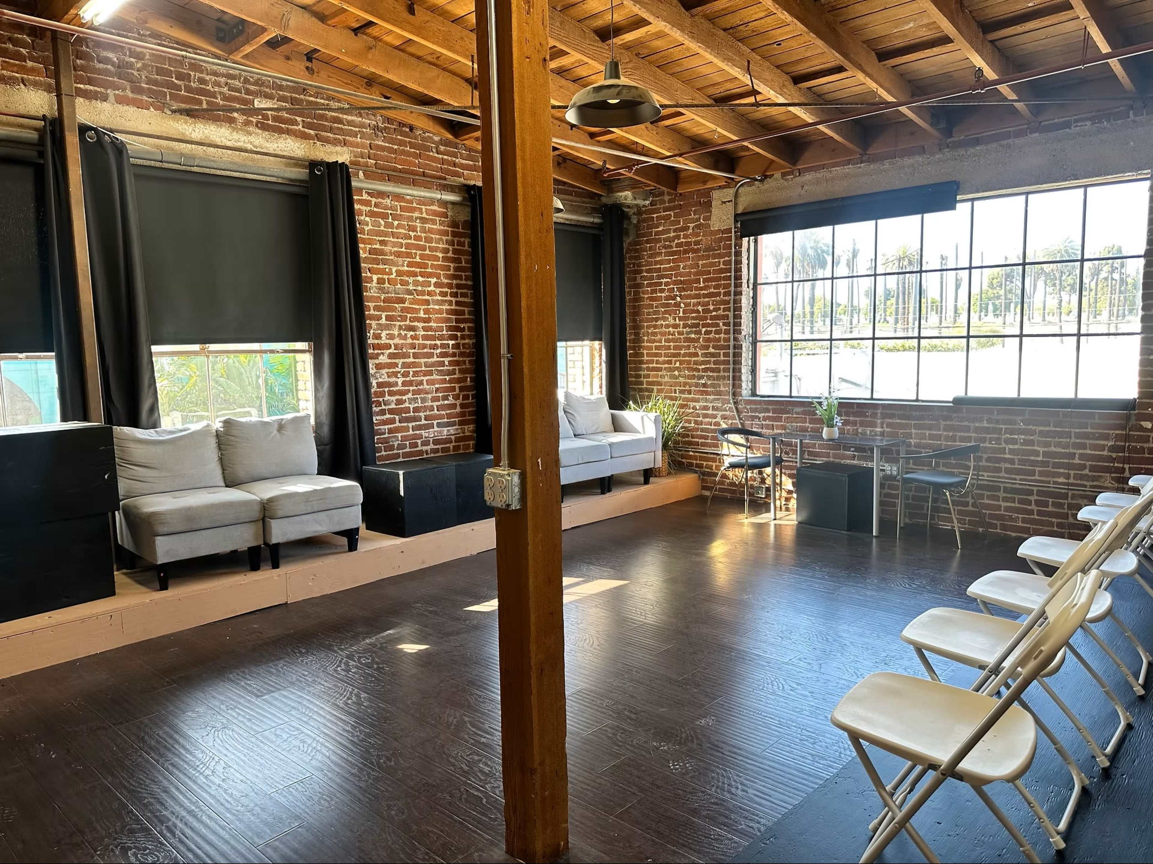 The image shows an empty, industrial-style room with exposed brick walls, wooden beams, and seating arranged in rows facing a large window.