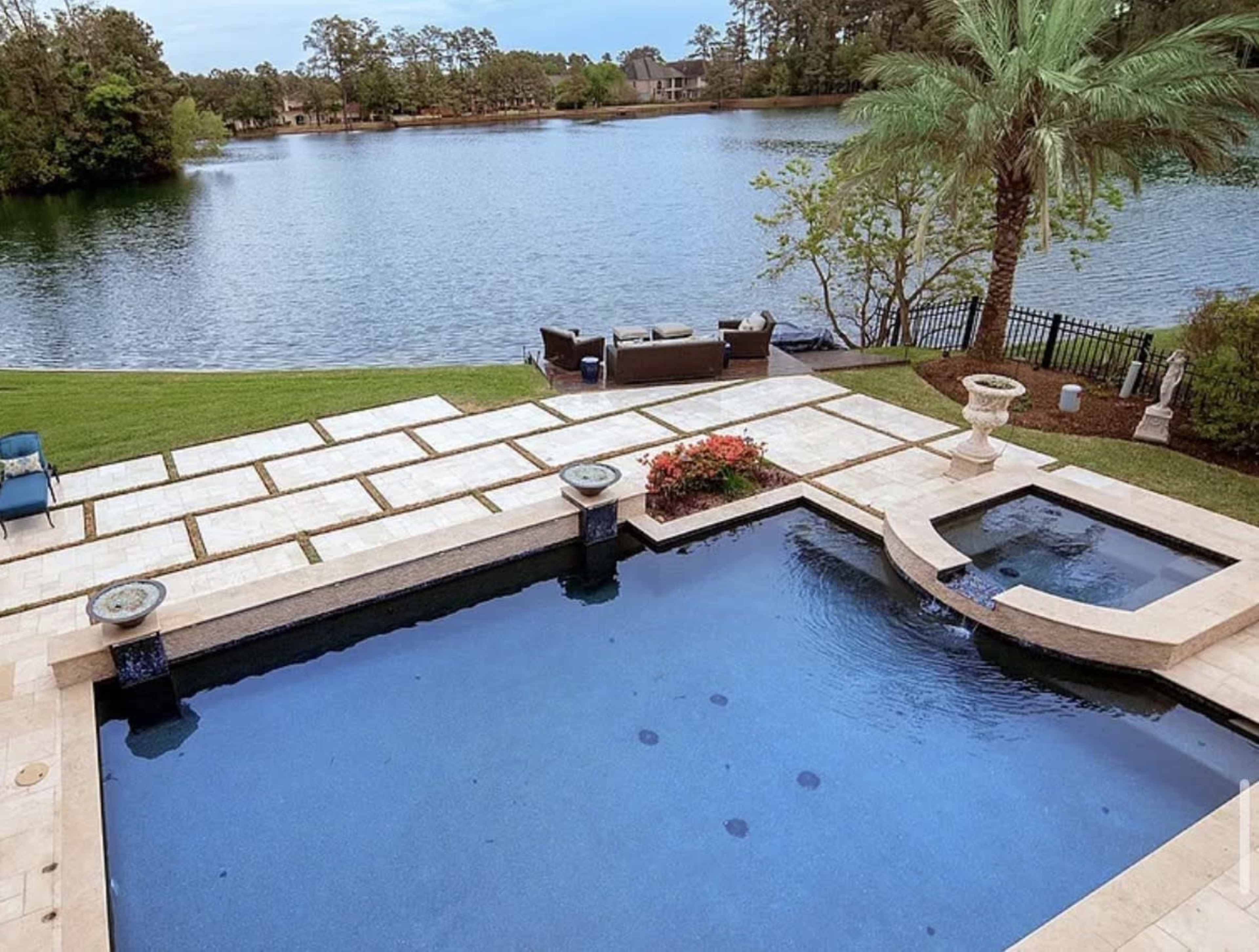 The image shows a landscaped backyard featuring a swimming pool, hot tub, patio furniture, and a view of a lake surrounded by trees.