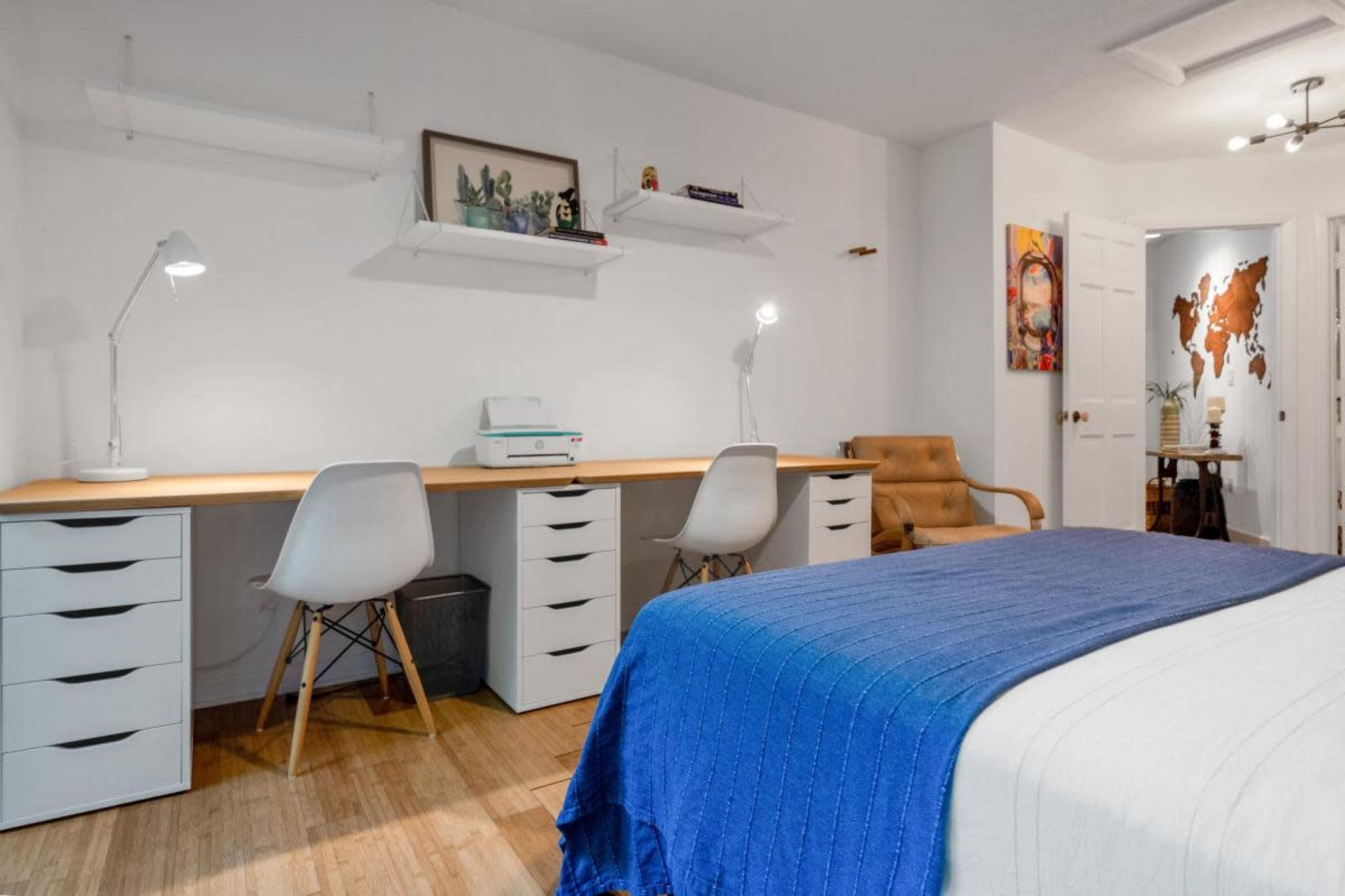 A modern bedroom featuring a bed with a blue blanket, a desk area with two white chairs, and wall shelves, along with a map artwork on one wall.