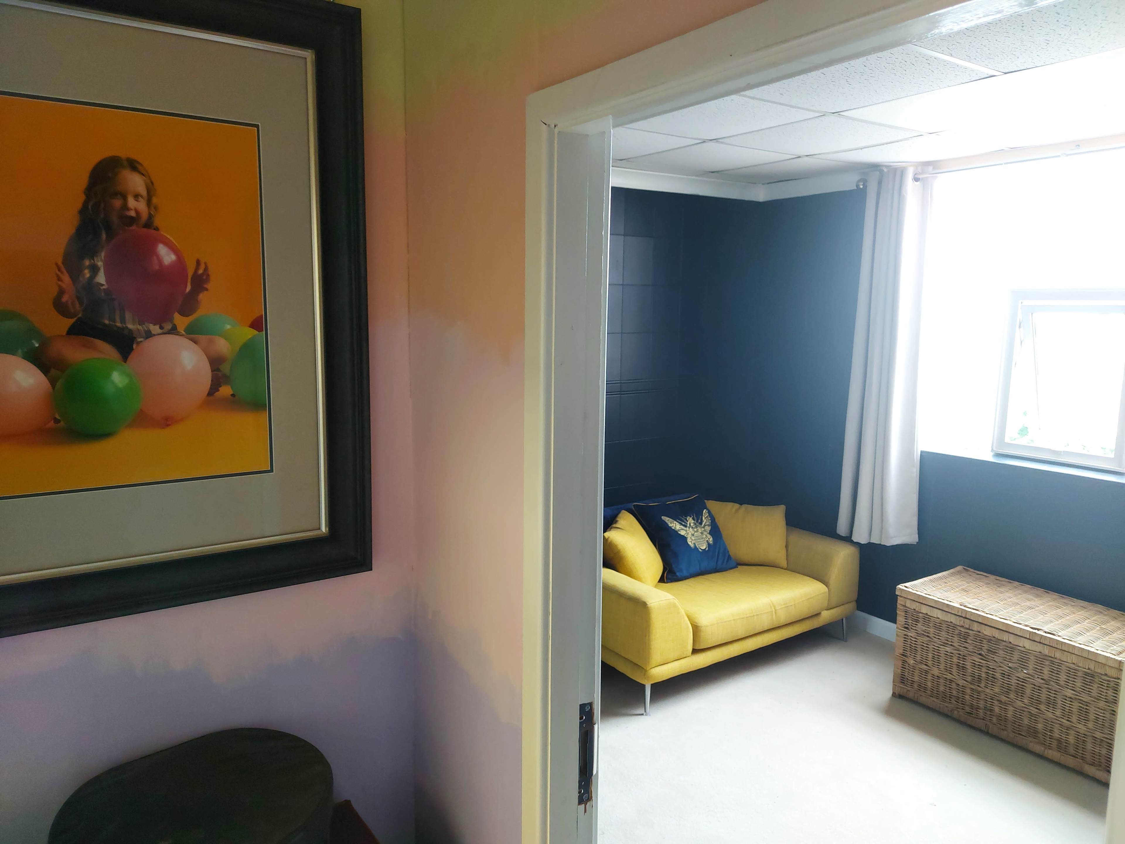 A brightly colored room with a yellow couch, a wicker storage box, and a large framed photograph of a child playing with colorful balloons.