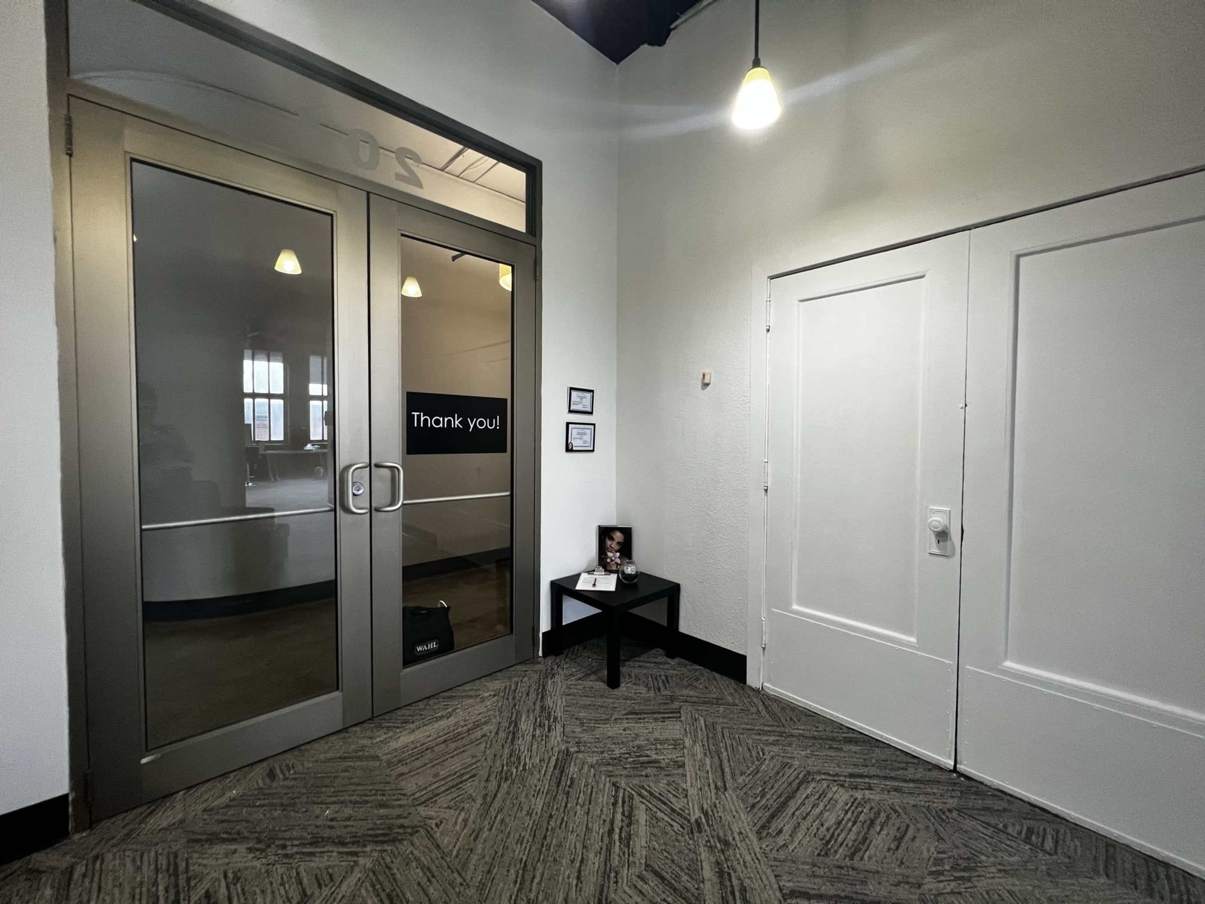 A corner of an indoor space featuring double glass doors labeled "Thank you," a small table with a framed photo, and a white closet door on the wall.