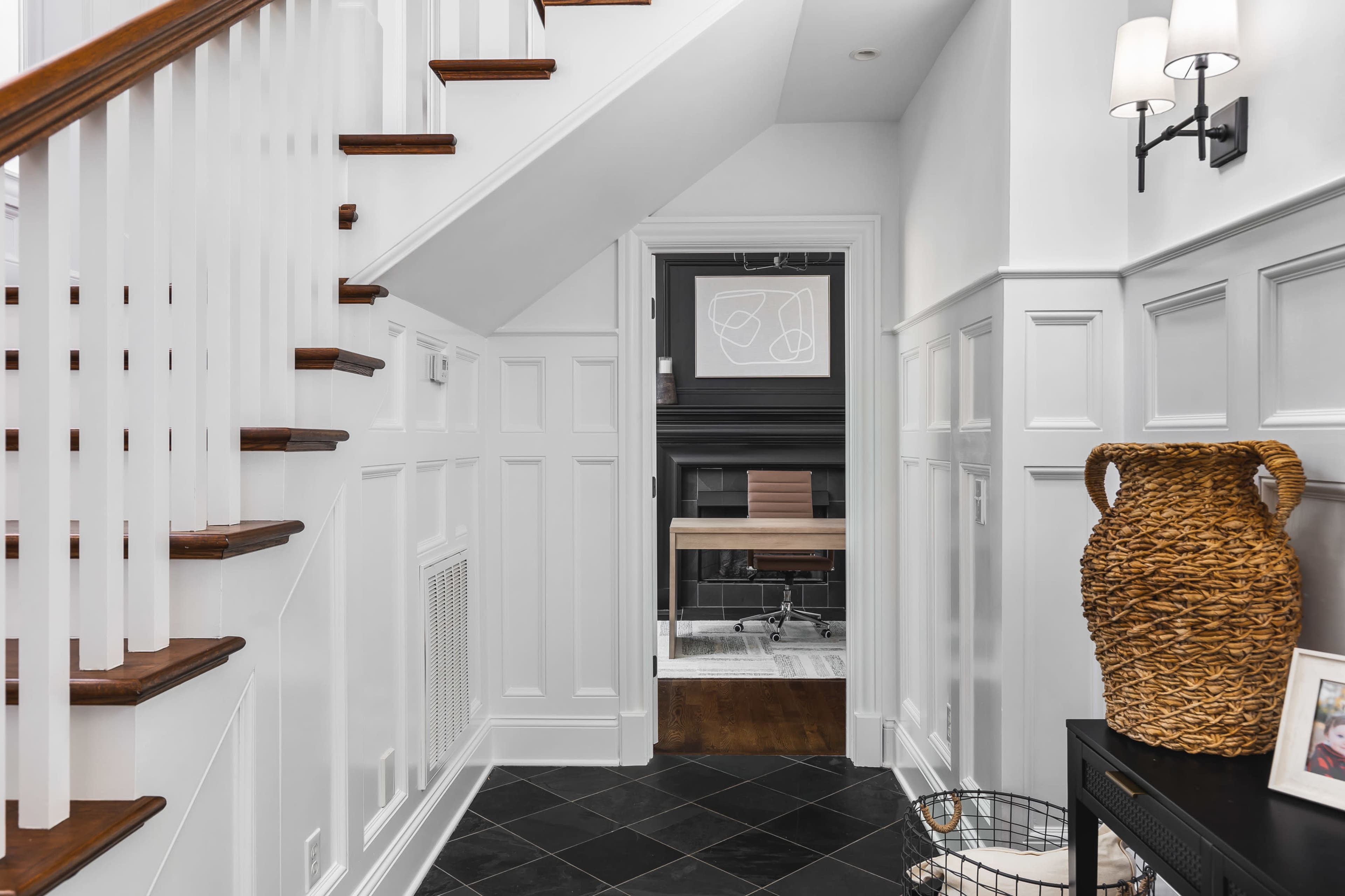 The image shows a well-lit hallway featuring a staircase, white paneling, and a view into a room with a black fireplace and modern decor.