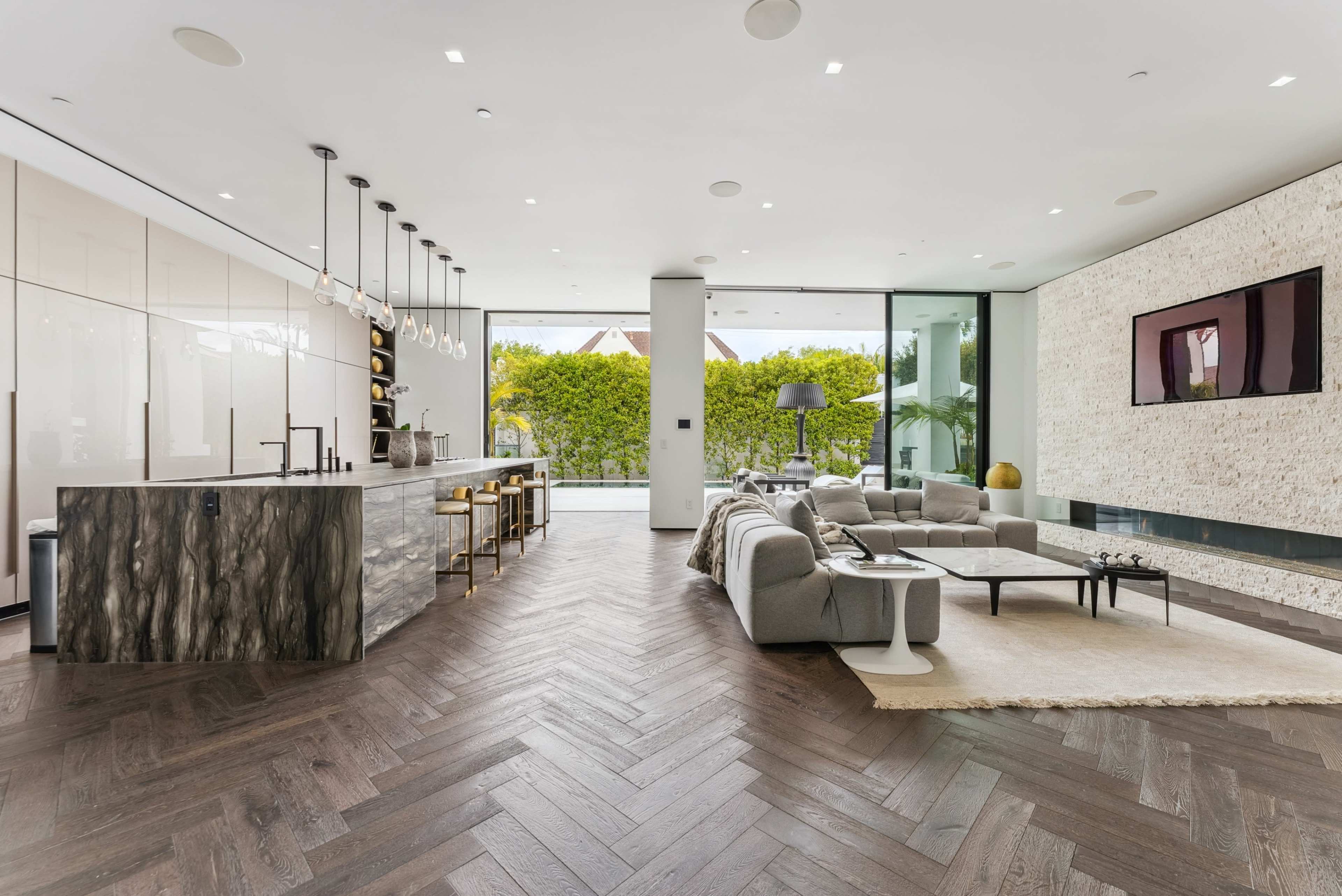 The image shows a modern living space featuring an open floor plan with a kitchen bar, a seating area, and large windows overlooking a green outdoor area.