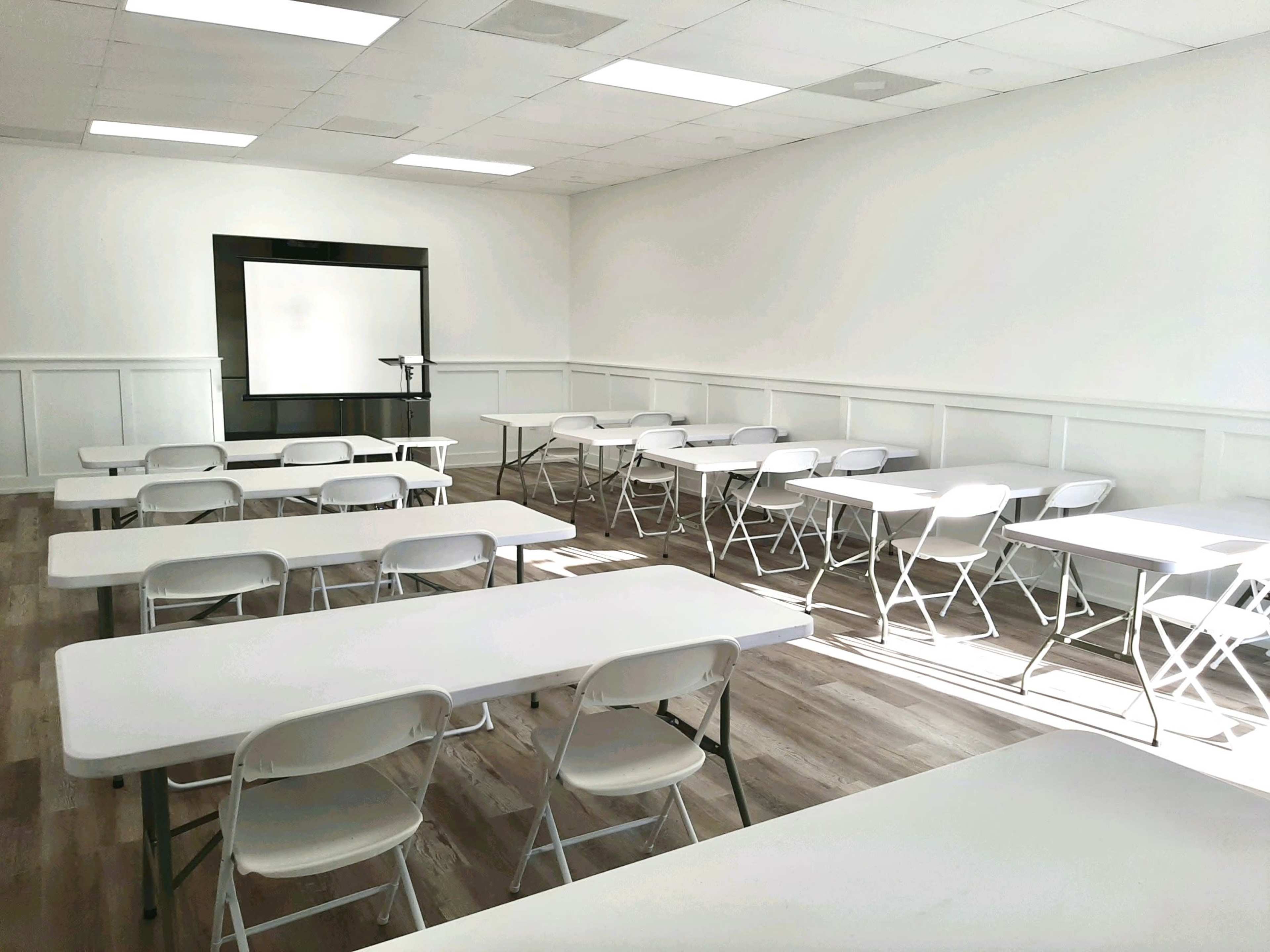 A brightly lit classroom features several rows of folding tables and chairs, with a projector screen mounted on the wall at the front.