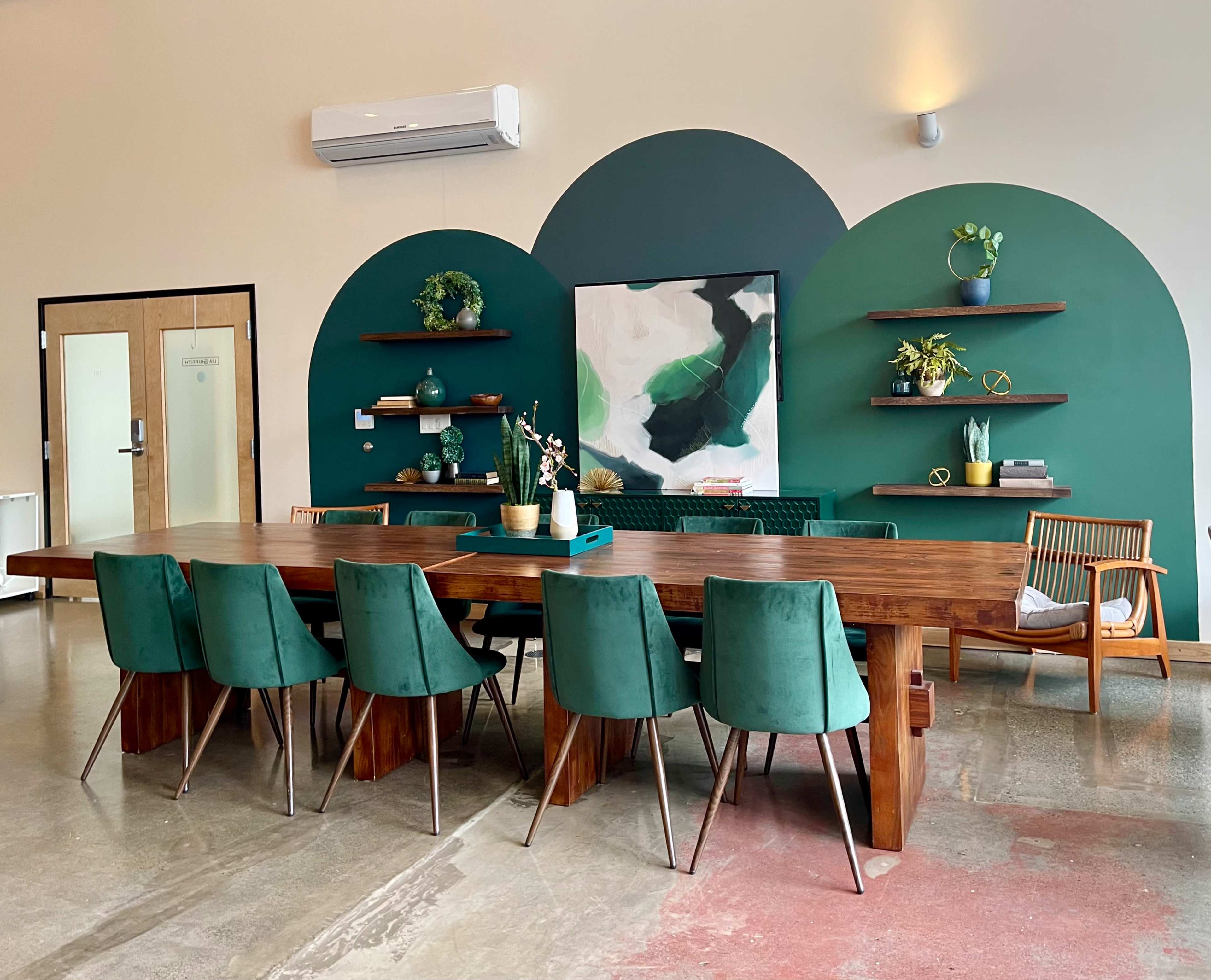 A wooden dining table with green chairs is set against a wall featuring two large green arches and shelves displaying decorative items.