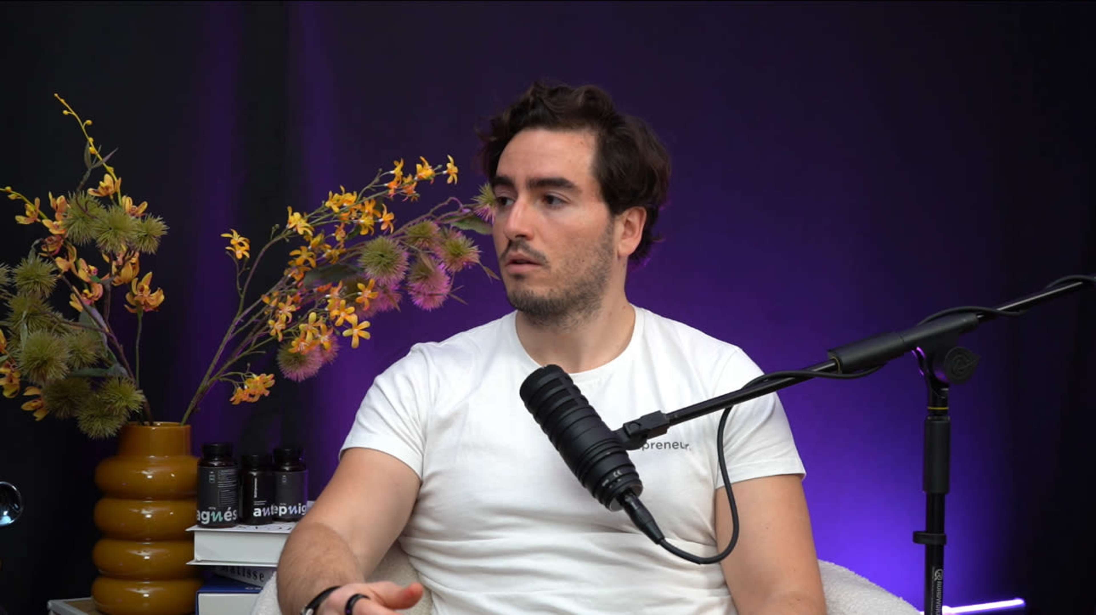 A man with dark hair, wearing a white t-shirt, is speaking while seated in front of a microphone and a plant arrangement against a dark backdrop.
