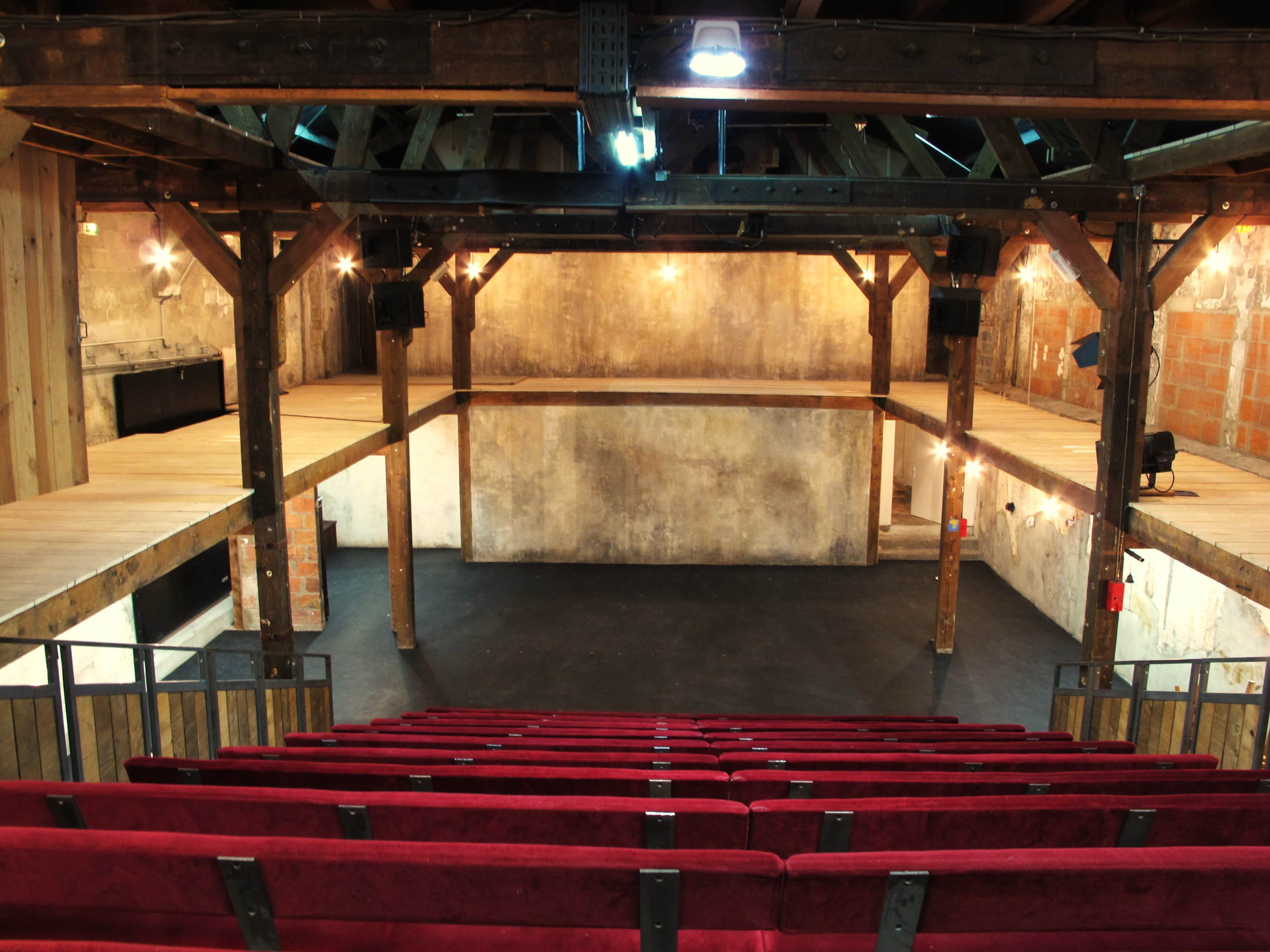 An empty theater with red seating facing a performance stage, featuring exposed wooden beams and a rustic interior.