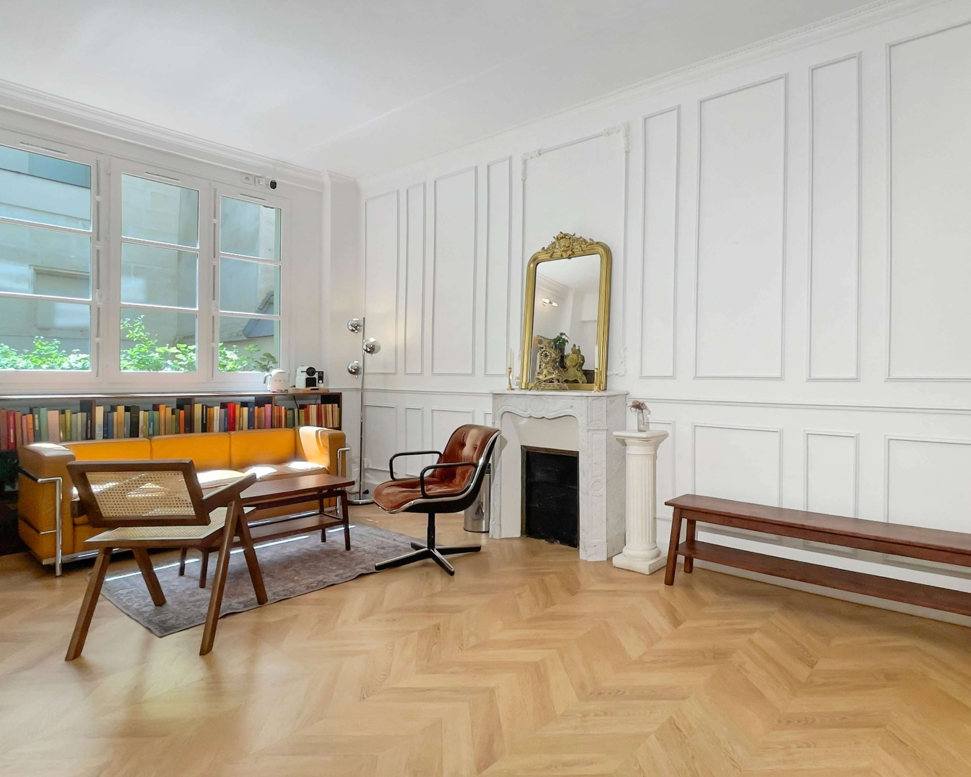 The image shows a well-lit living room featuring a yellow sofa, a bookshelf, a fireplace with a mirror above it, and wooden furniture on a herringbone-patterned floor.