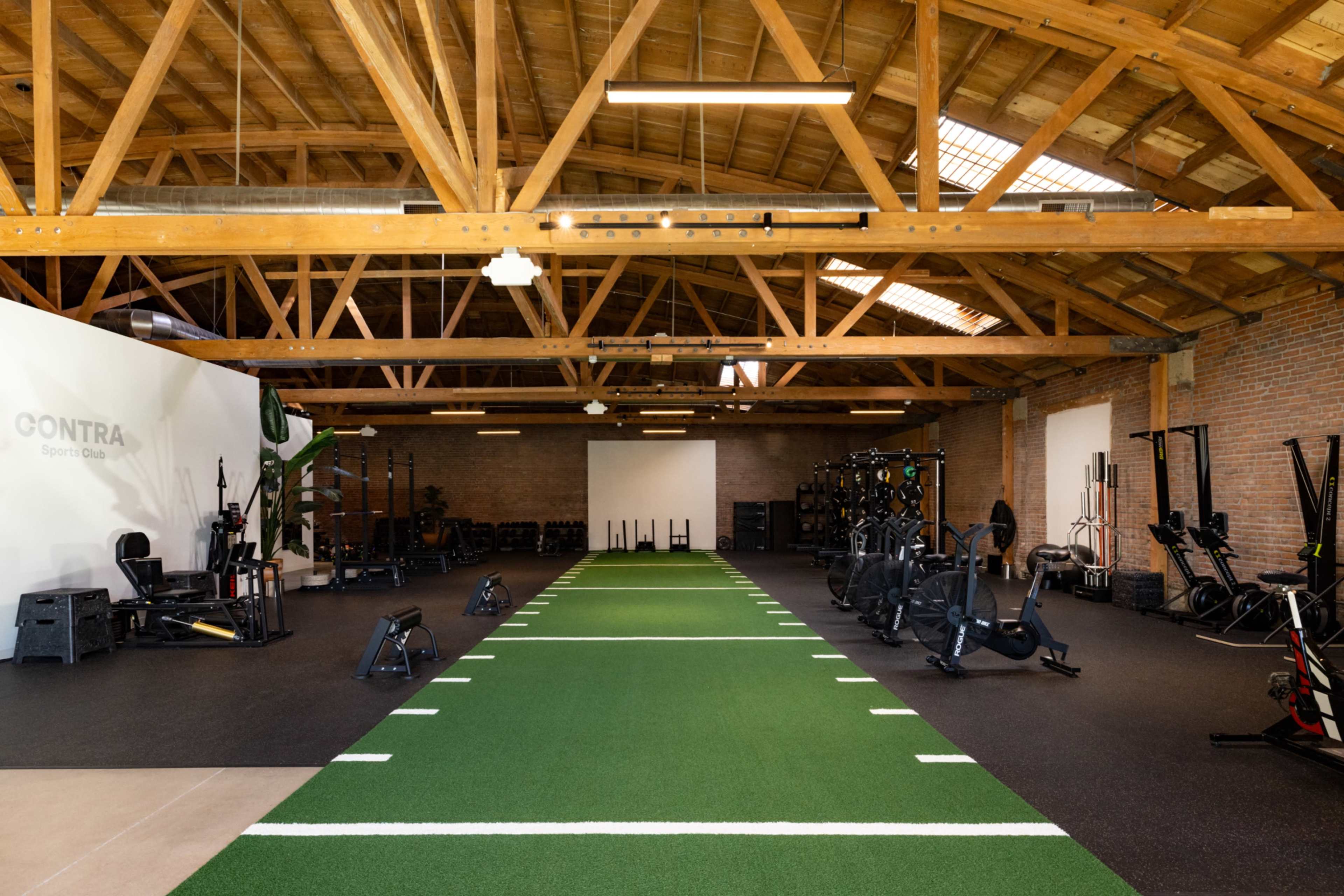 The image shows a spacious gym with a green turf section in the center, surrounded by various fitness equipment on both sides.