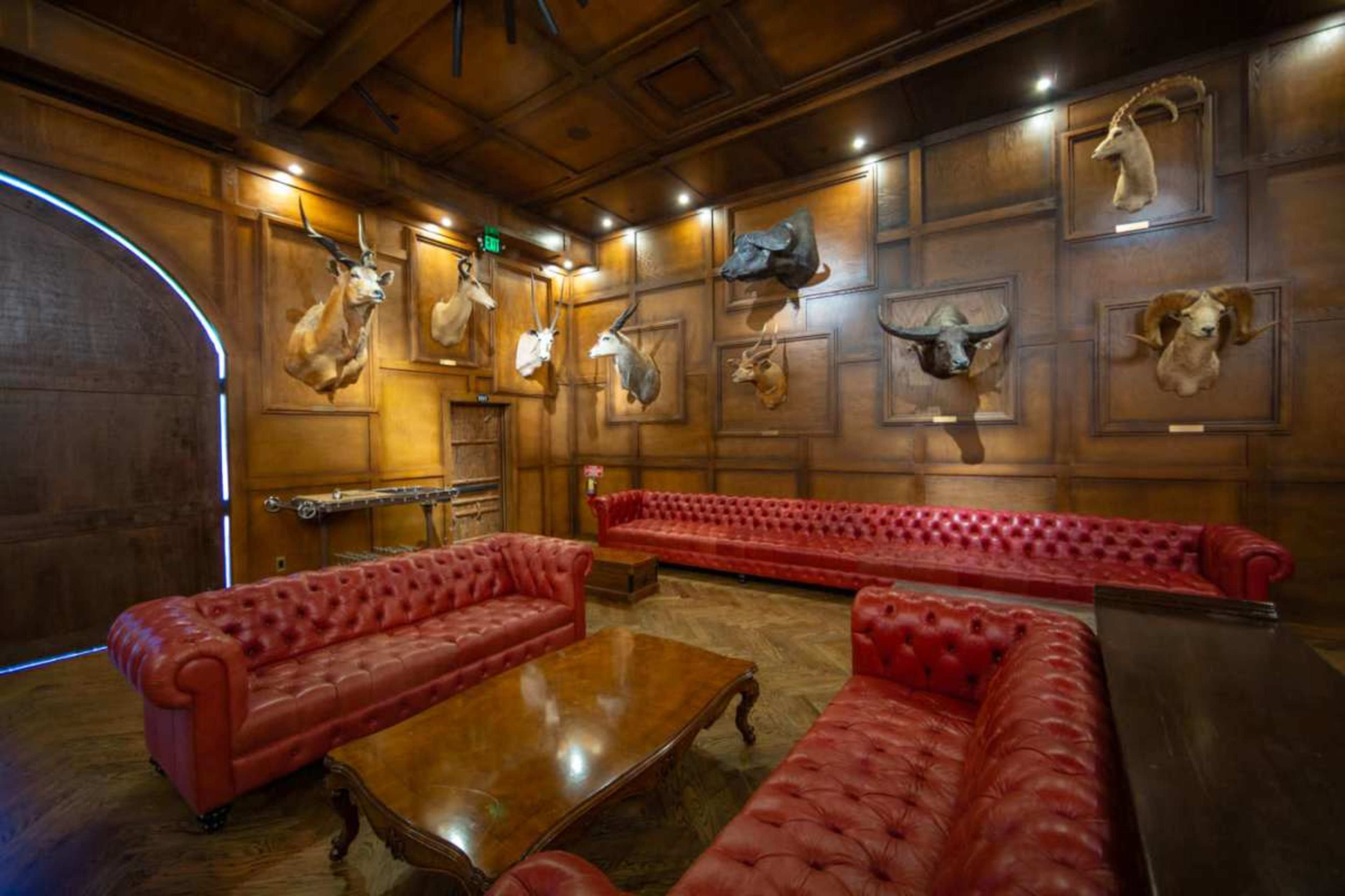 The image shows a room with dark wood paneling, featuring two red leather chesterfield sofas and several mounted animal heads on the walls.