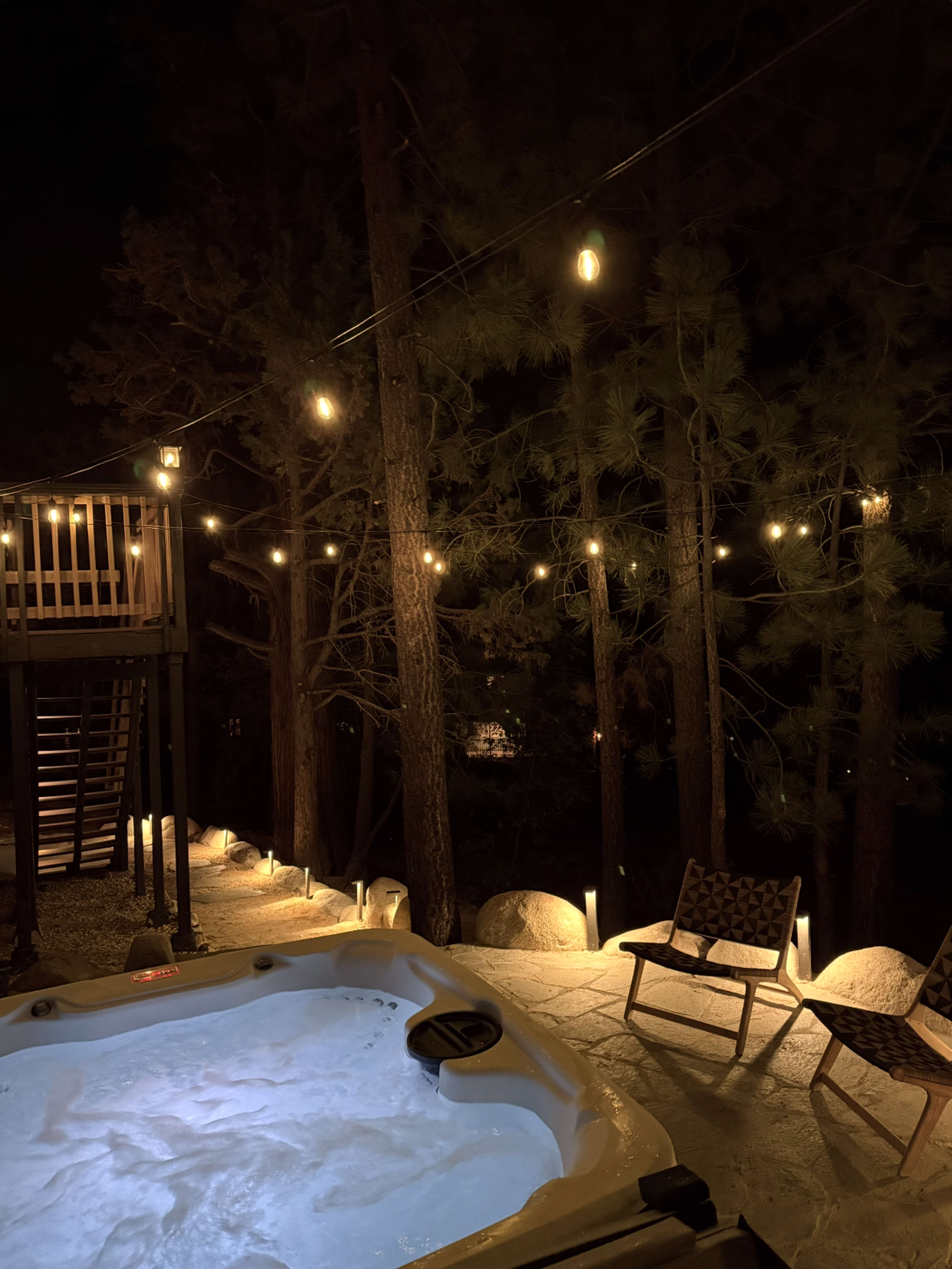 A hot tub filled with water sits beside a tree-lined area illuminated by string lights at night.