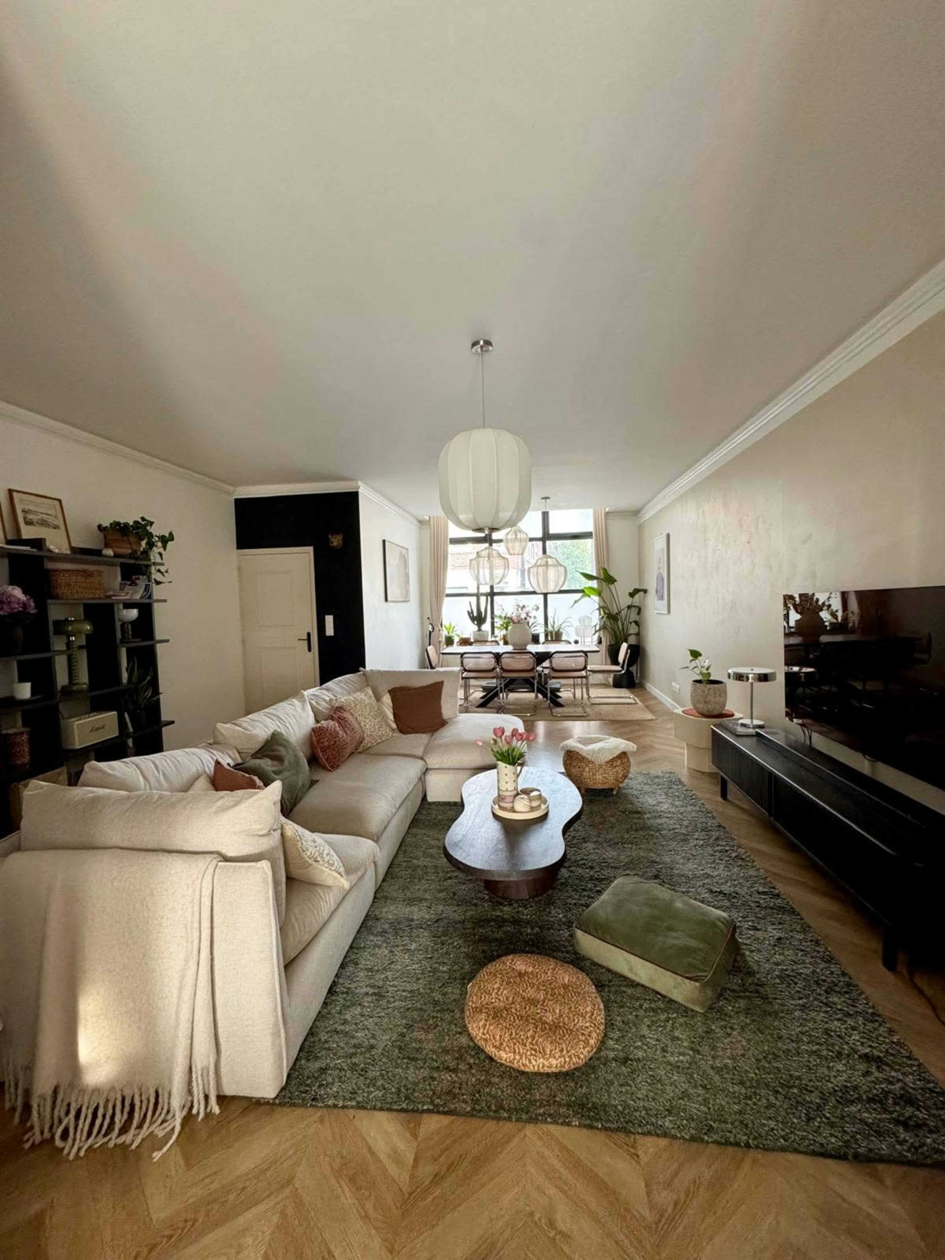 A spacious living room features a large sectional sofa, a wooden coffee table, and a television set, with plants and decorative items arranged around the space.