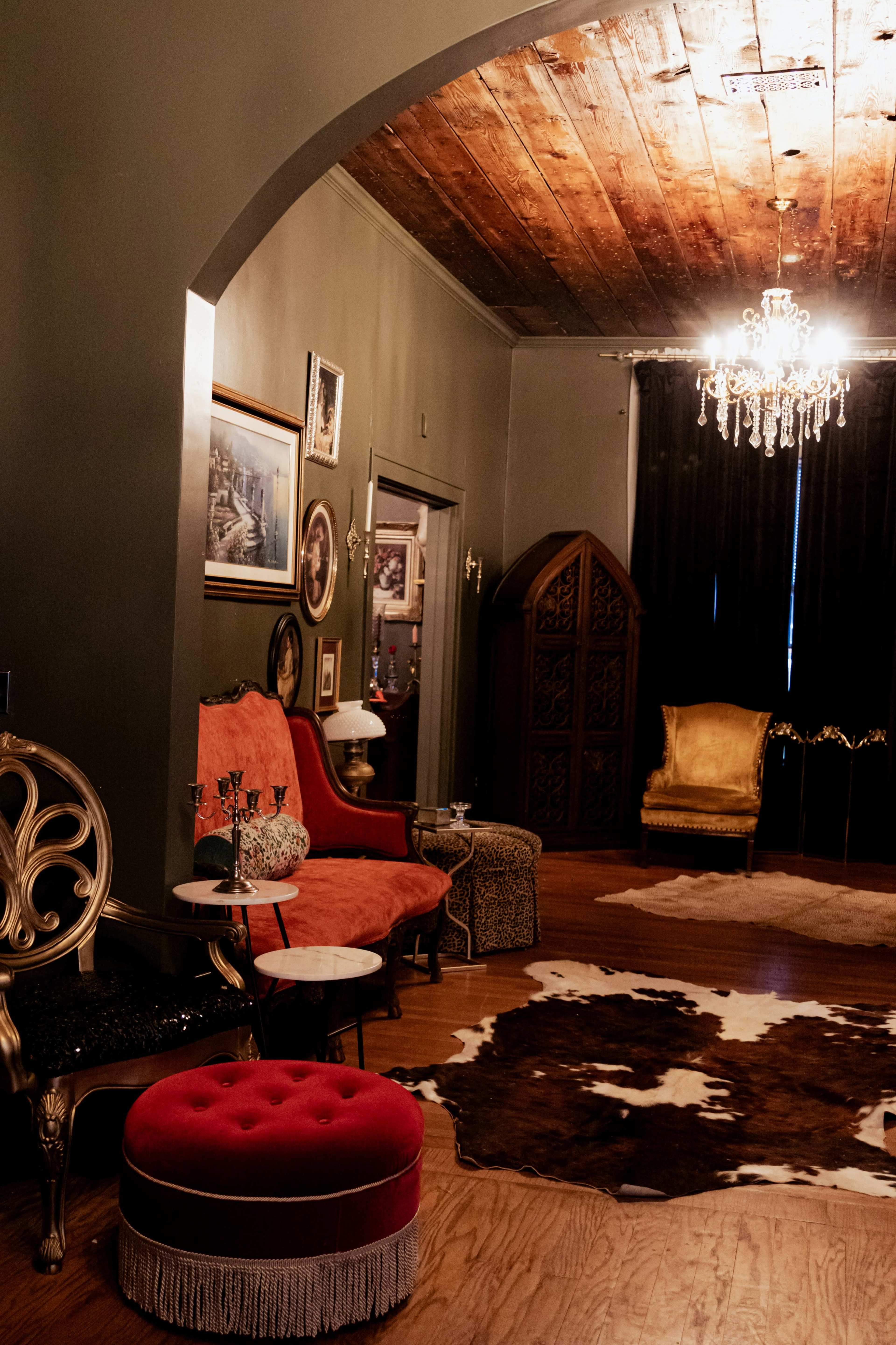 The image depicts a stylishly decorated room featuring a mix of vintage furniture, including a red velvet chair, a golden armchair, and a cowhide rug on the wooden floor.