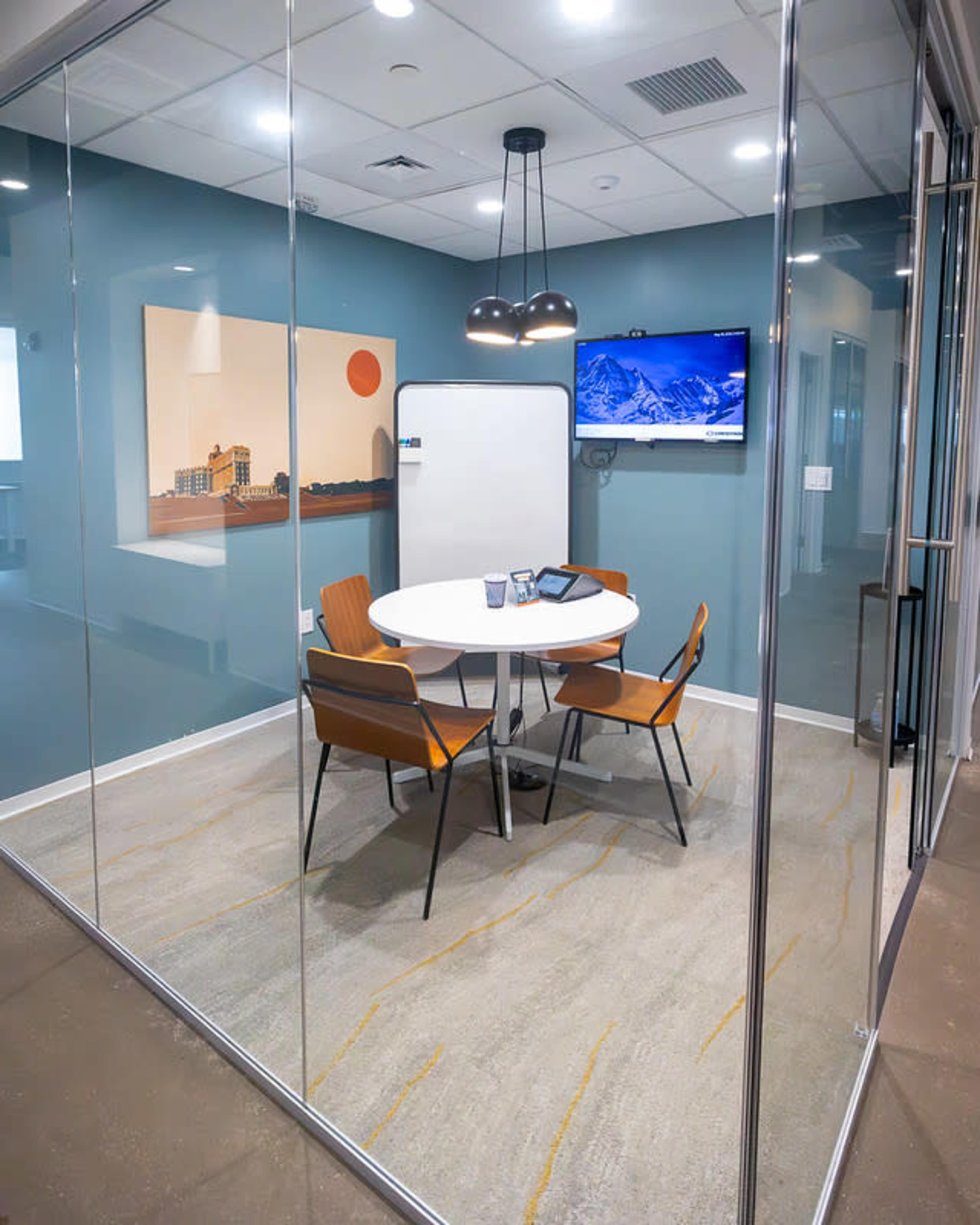 The image shows a modern conference room with a circular table, four chairs, a whiteboard, and a television mounted on the wall, all enclosed by glass walls.