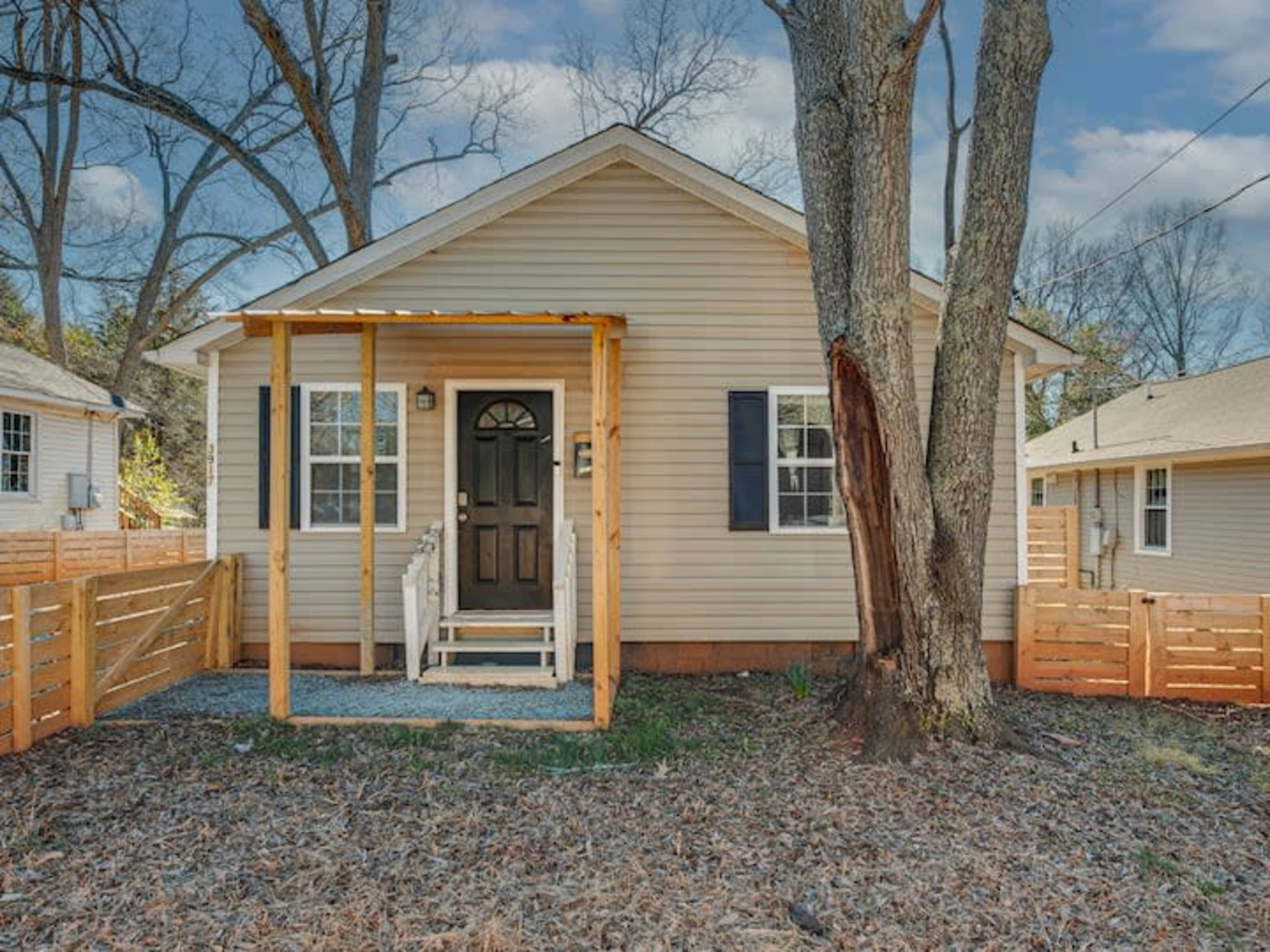 A small house with a front porch and wooden steps is surrounded by a fenced yard and trees.