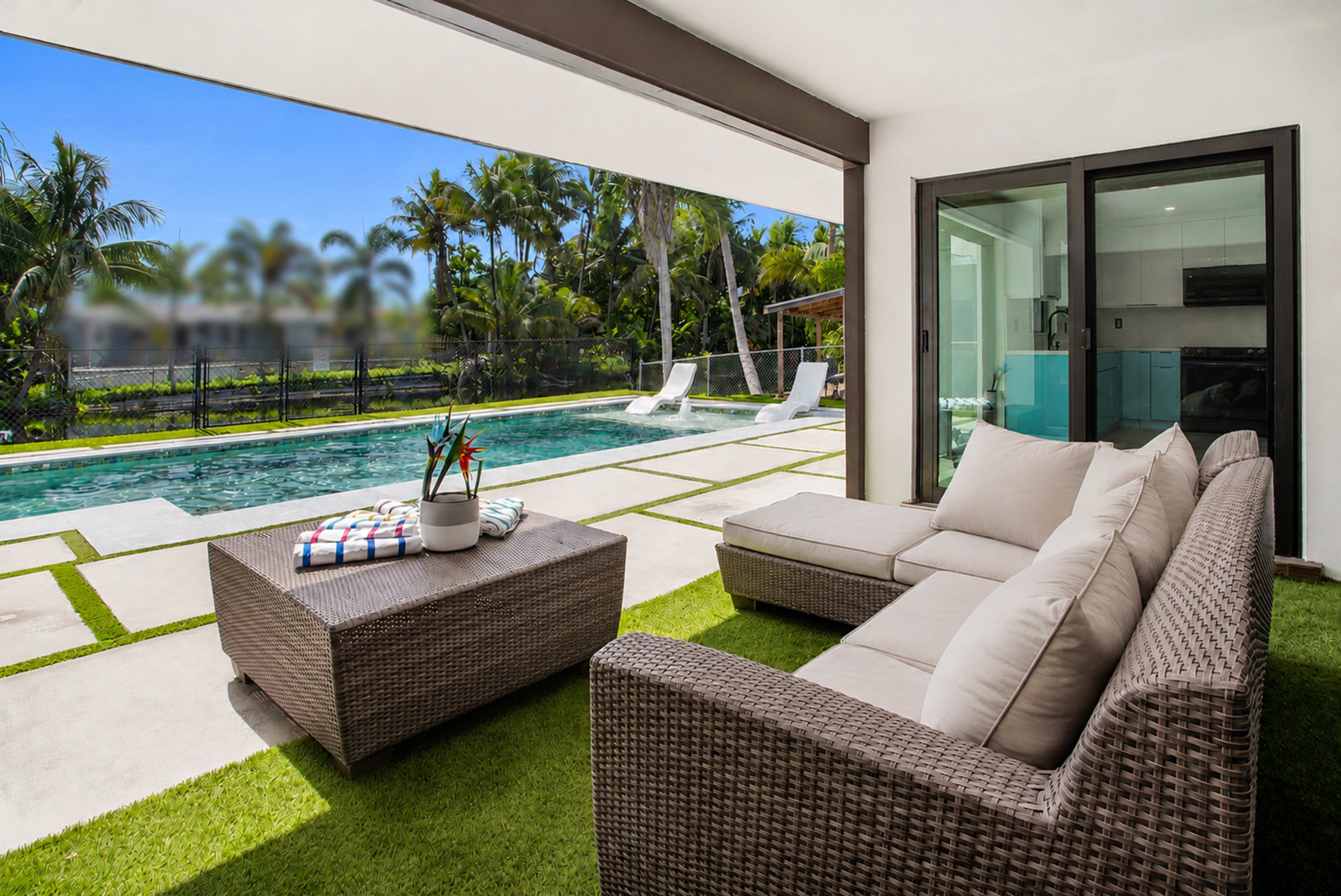 A modern outdoor seating area with a wicker sofa and coffee table overlooks a swimming pool surrounded by palm trees.