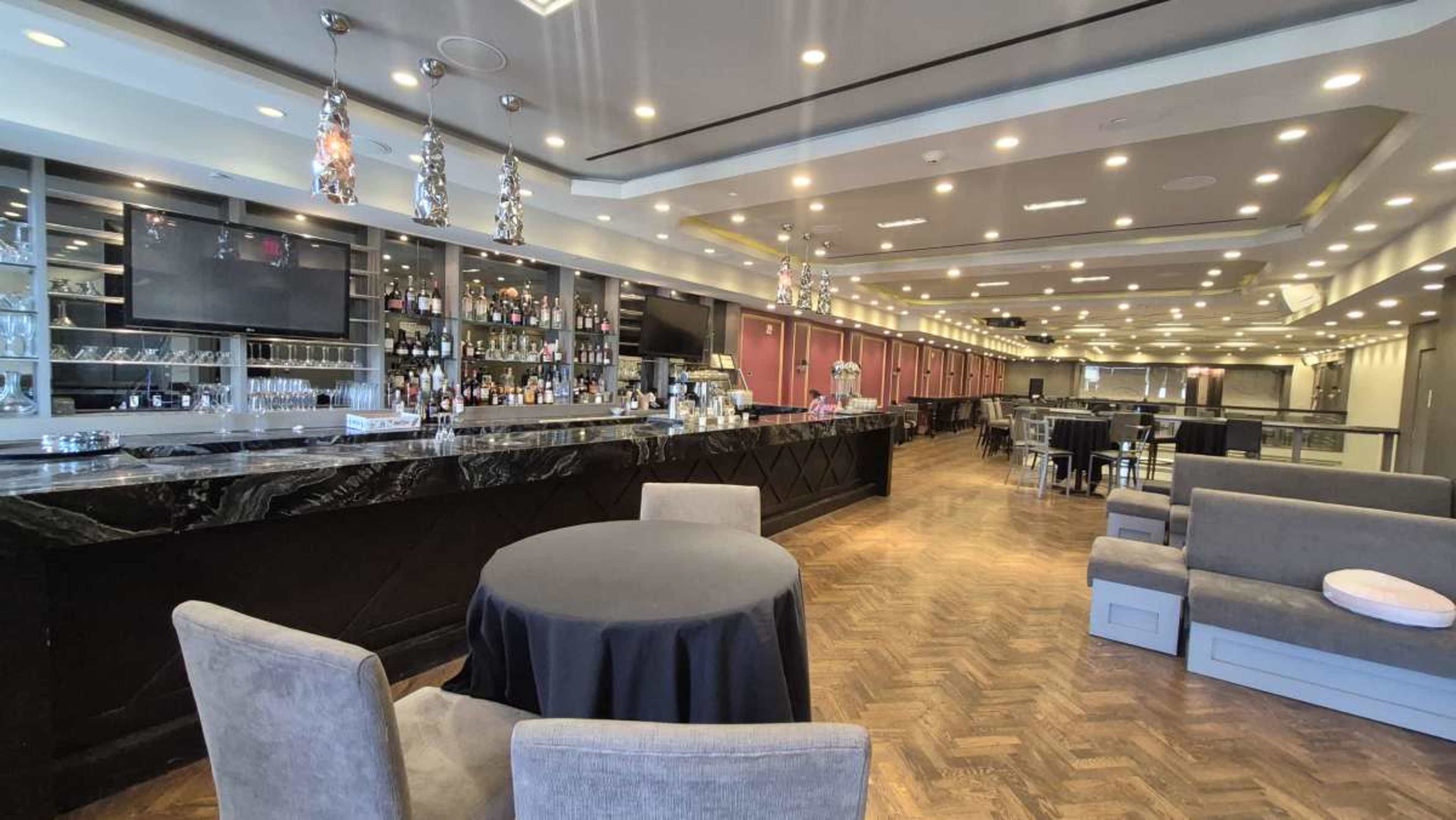 The image shows a spacious, well-lit bar area with a long counter, shelves of drinks, and seating arranged along the sides.