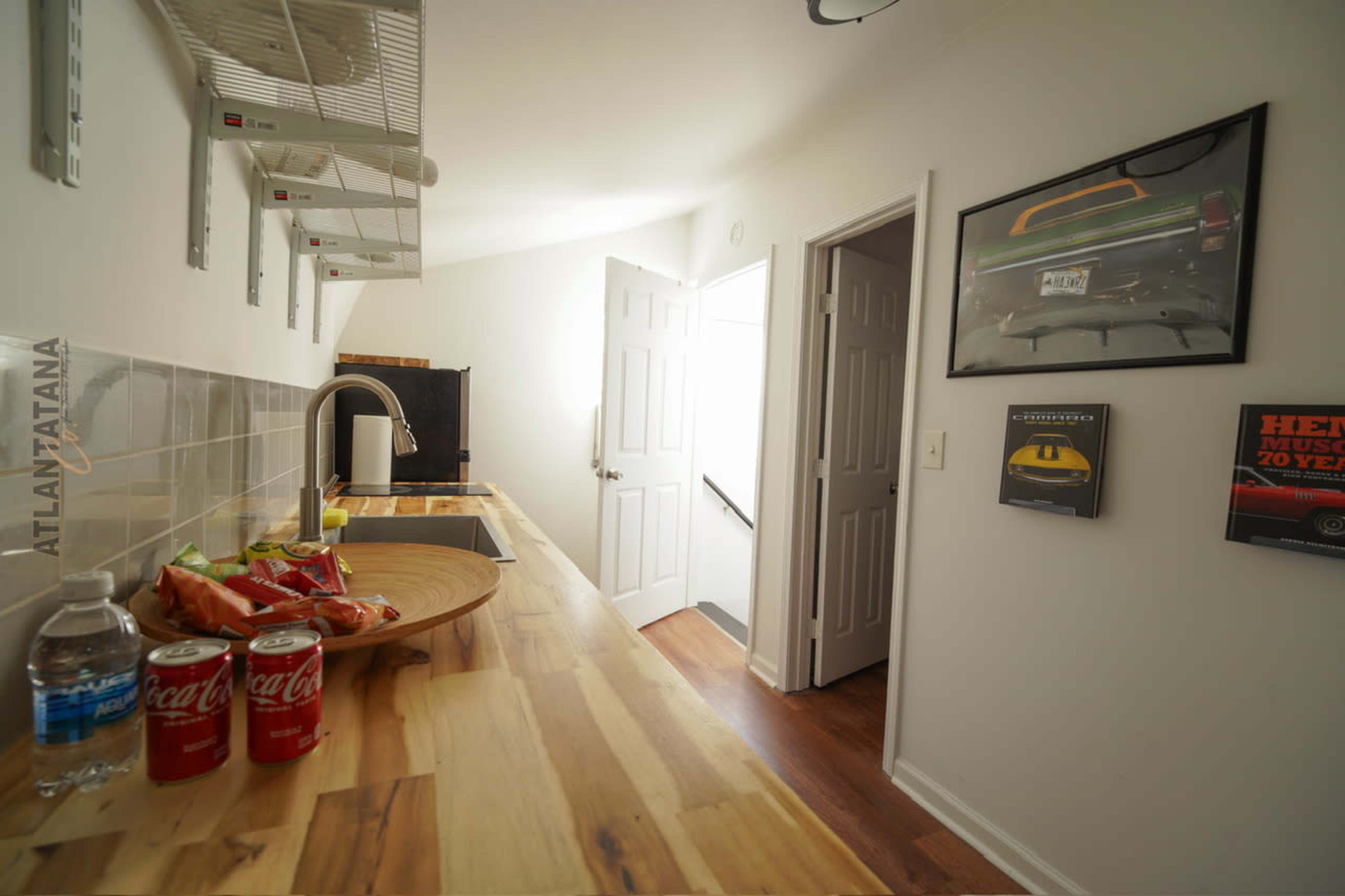 The image shows a narrow kitchen space featuring a wooden countertop, a sink, a shelf with appliances, and a door leading to another room.