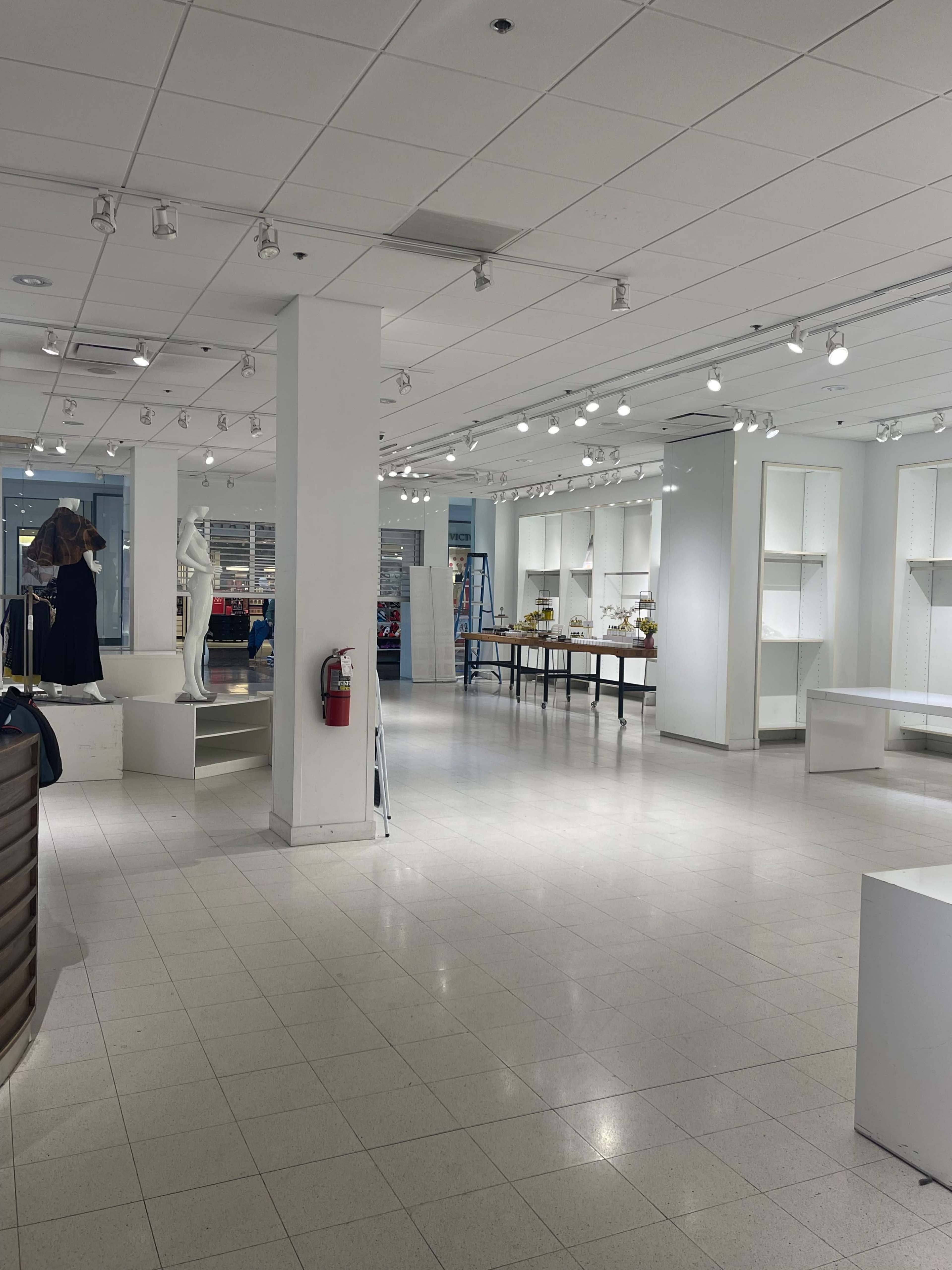 The image shows an empty retail space with white walls, bright lighting, a statue, and a ladder, indicating preparation for a display or setup.