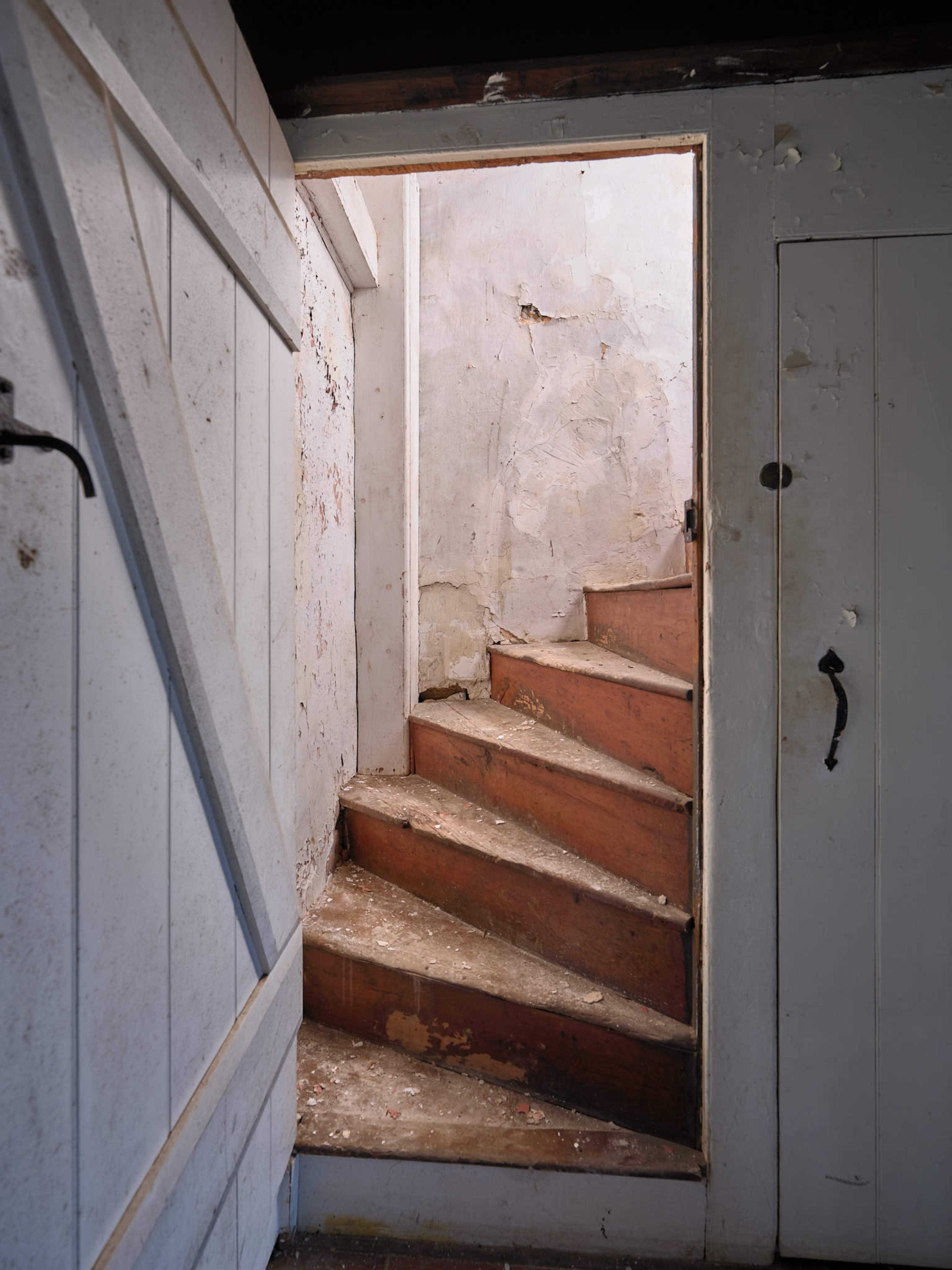 A wooden staircase leads upwards from a dimly lit area, flanked by peeling white walls and a closed door on the right.
