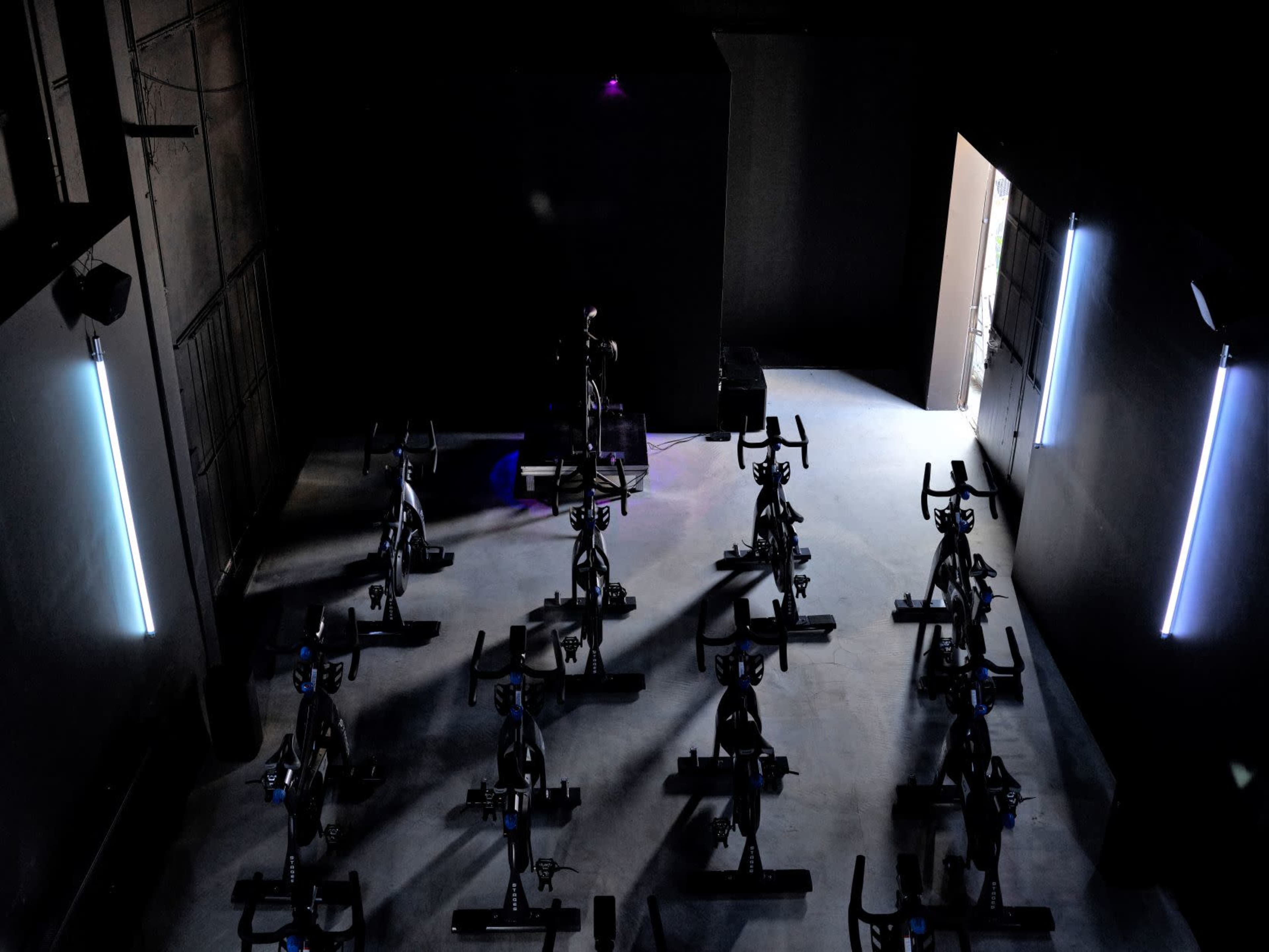 A dimly lit indoor cycling studio features multiple stationary bikes arranged in rows, with an entrance visible in the background.