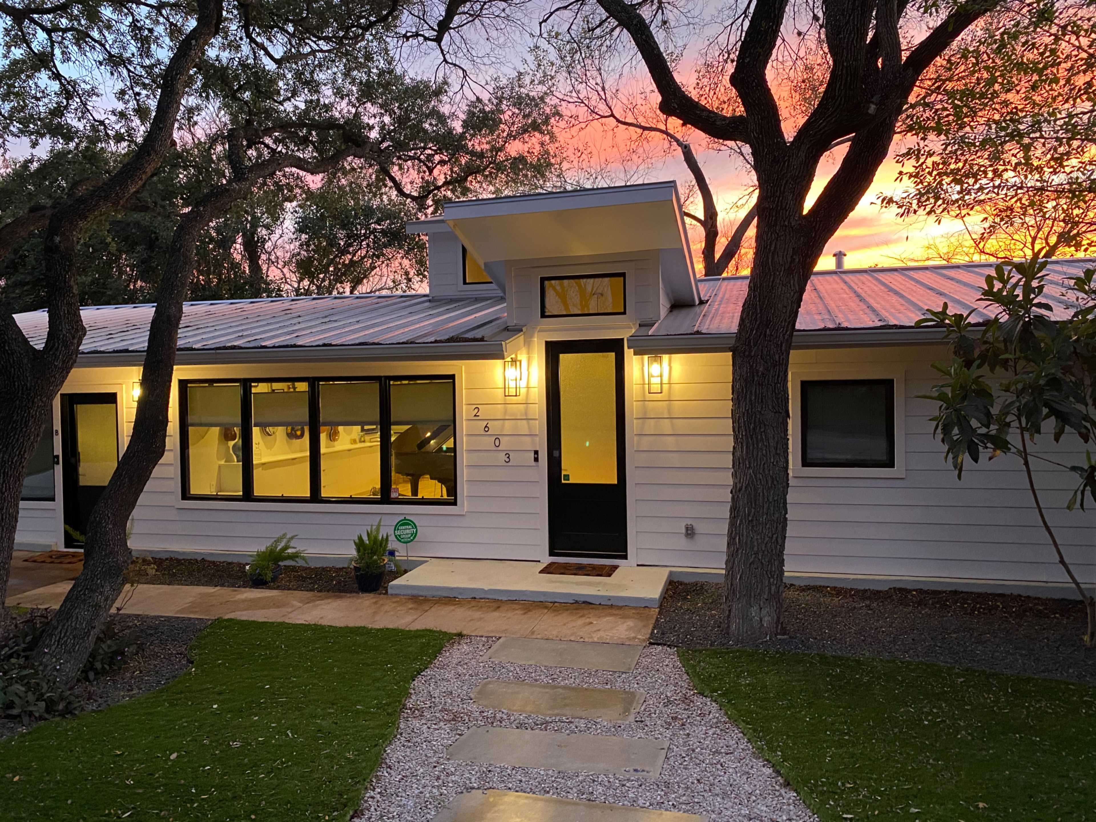 A modern white house with a metal roof and large windows is illuminated at dusk, surrounded by trees and a stone path leading to its entrance.