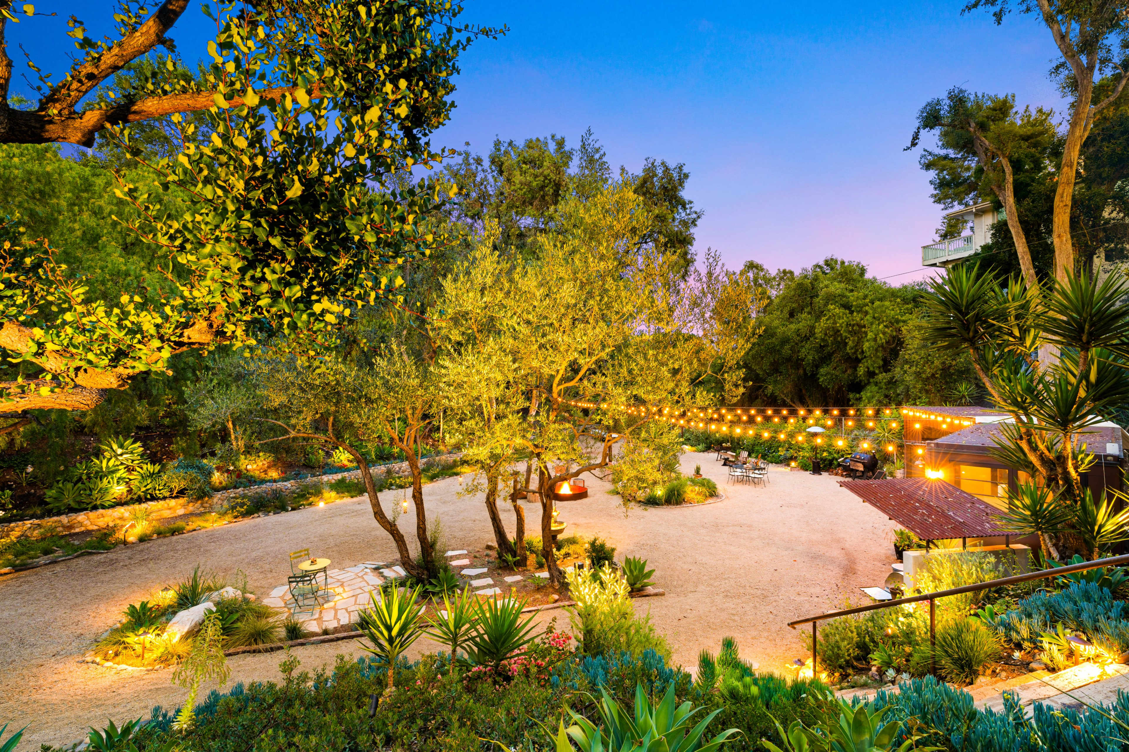 A landscaped outdoor area features gravel paths, trees, and string lights, creating a serene space for gathering.