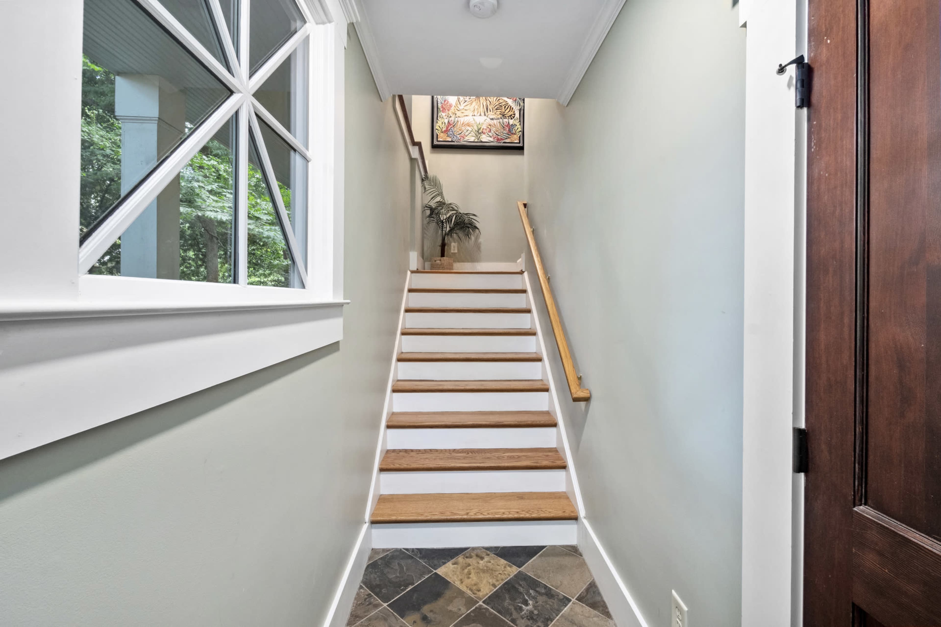 A staircase leading upward, flanked by a window on one side and a wooden door on the other, with a small plant placed at the top.